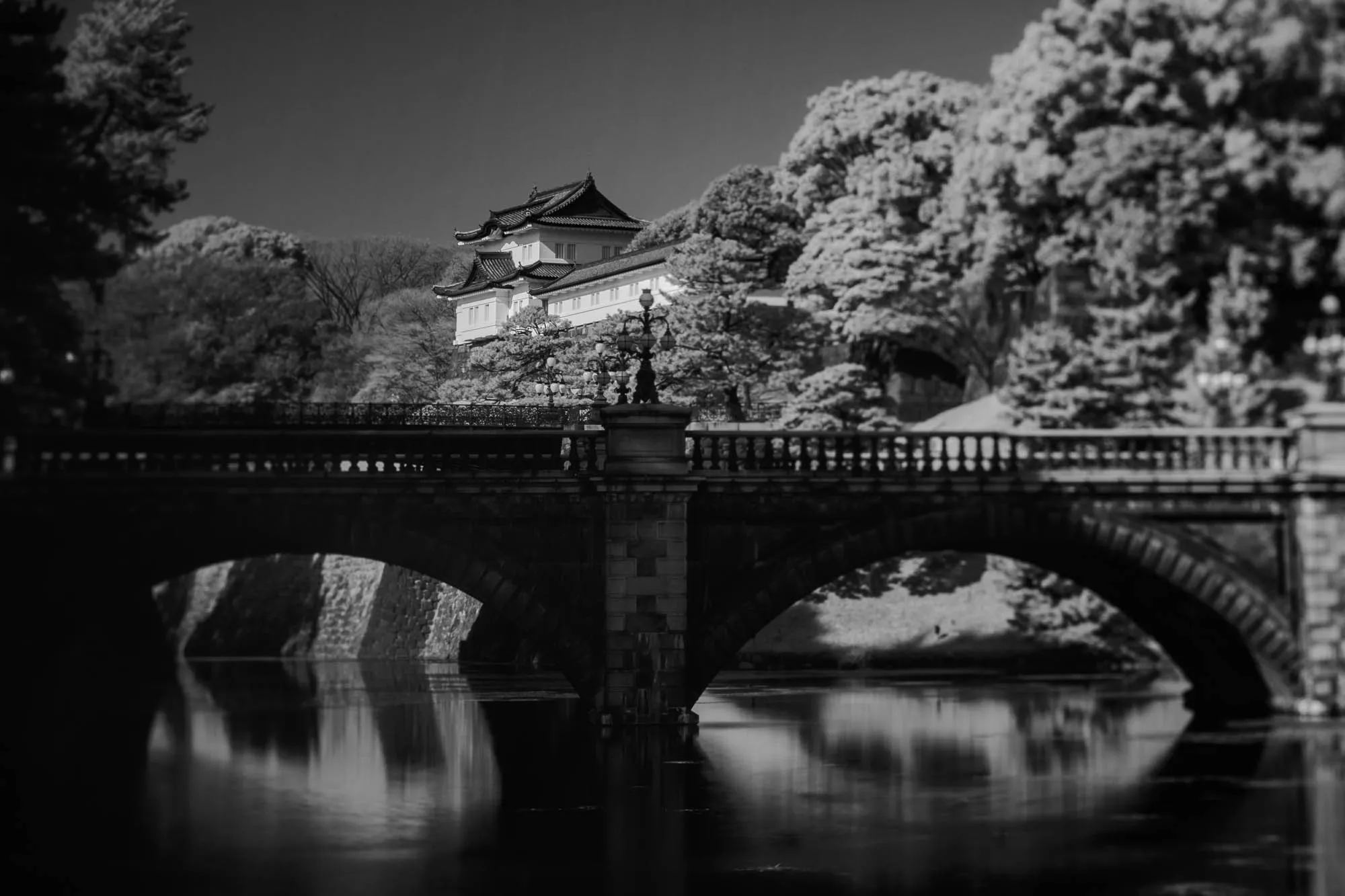 The image shows a stone bridge over a calm body of water. The bridge has two arches and is made of grey stone. It has a railing along both sides. The bridge is surrounded by trees and a stone wall. There is a white building on the other side of the bridge, and a few lampposts are seen on both sides of the bridge. The water is still and reflects the sky and the surrounding scenery. The image evokes a sense of tranquility and peace.