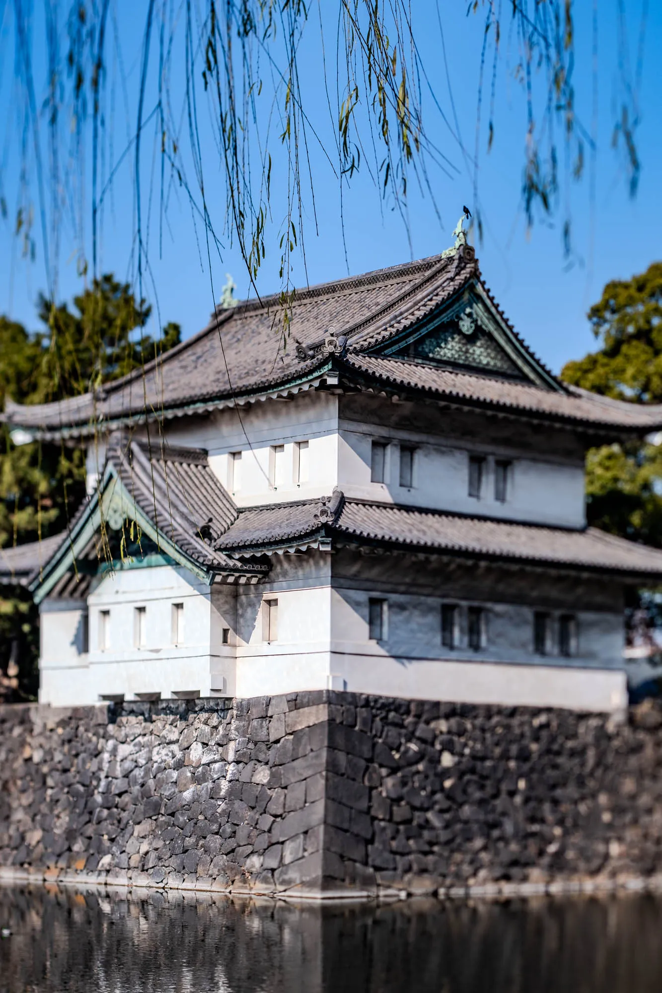 A Japanese style building with a grey tiled roof. The building is white with a black trim around the windows. There are three windows on each of the two floors. The building is situated on a large stone wall which is in front of a body of water. The image is framed by the long, thin branches of a weeping willow tree. The branches are in the foreground of the image. The sky is blue.