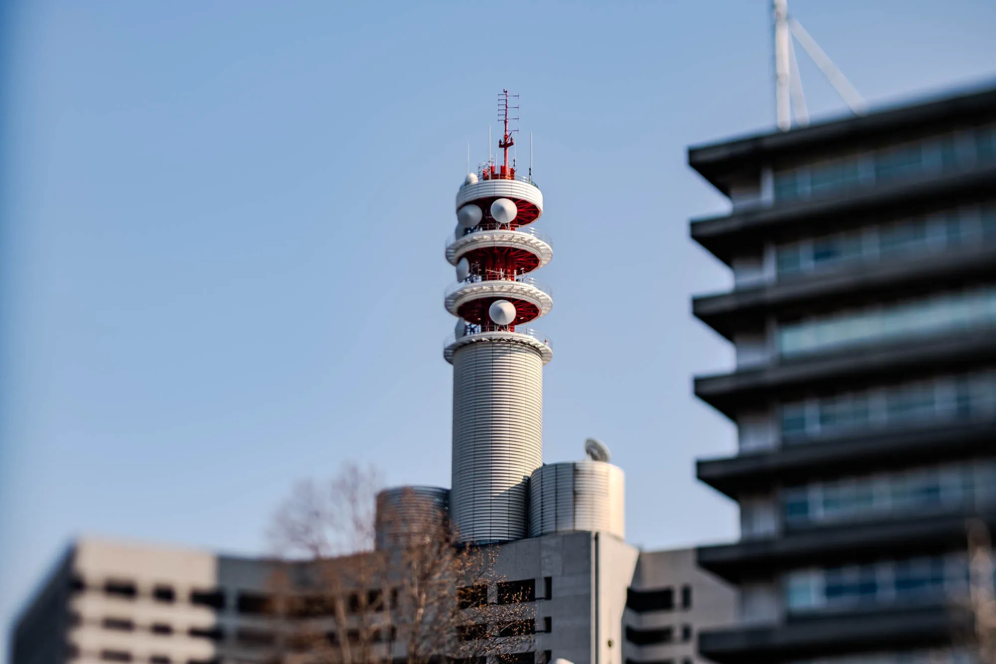 The image shows a tall, cylindrical communication tower with a red and white structure at the top, with multiple satellite dishes.  The tower is in front of a large building with several horizontal rows of windows. The background is a clear blue sky.  A bare-branched tree is partially visible in the foreground.