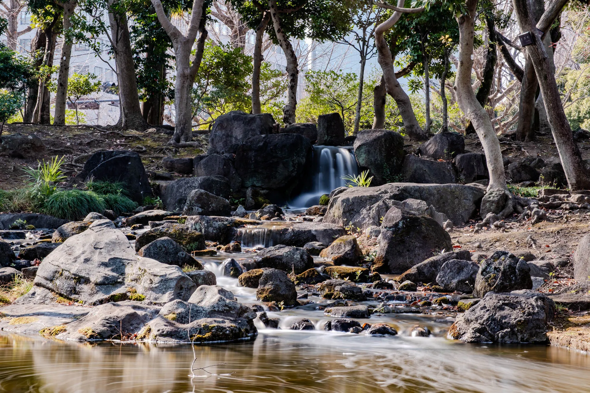 The image is a close-up of a small waterfall cascading down a rocky stream bed in a forested area. There are a few trees visible on either side of the waterfall, with their trunks and branches partially obscuring the view. The water in the stream is flowing quickly, creating a blurred effect. The rocks in the stream bed are grey and brown, and some are covered in moss. The water in the pond below the waterfall is still and reflective, reflecting the surrounding trees and sky. The overall impression is one of peace and tranquility.