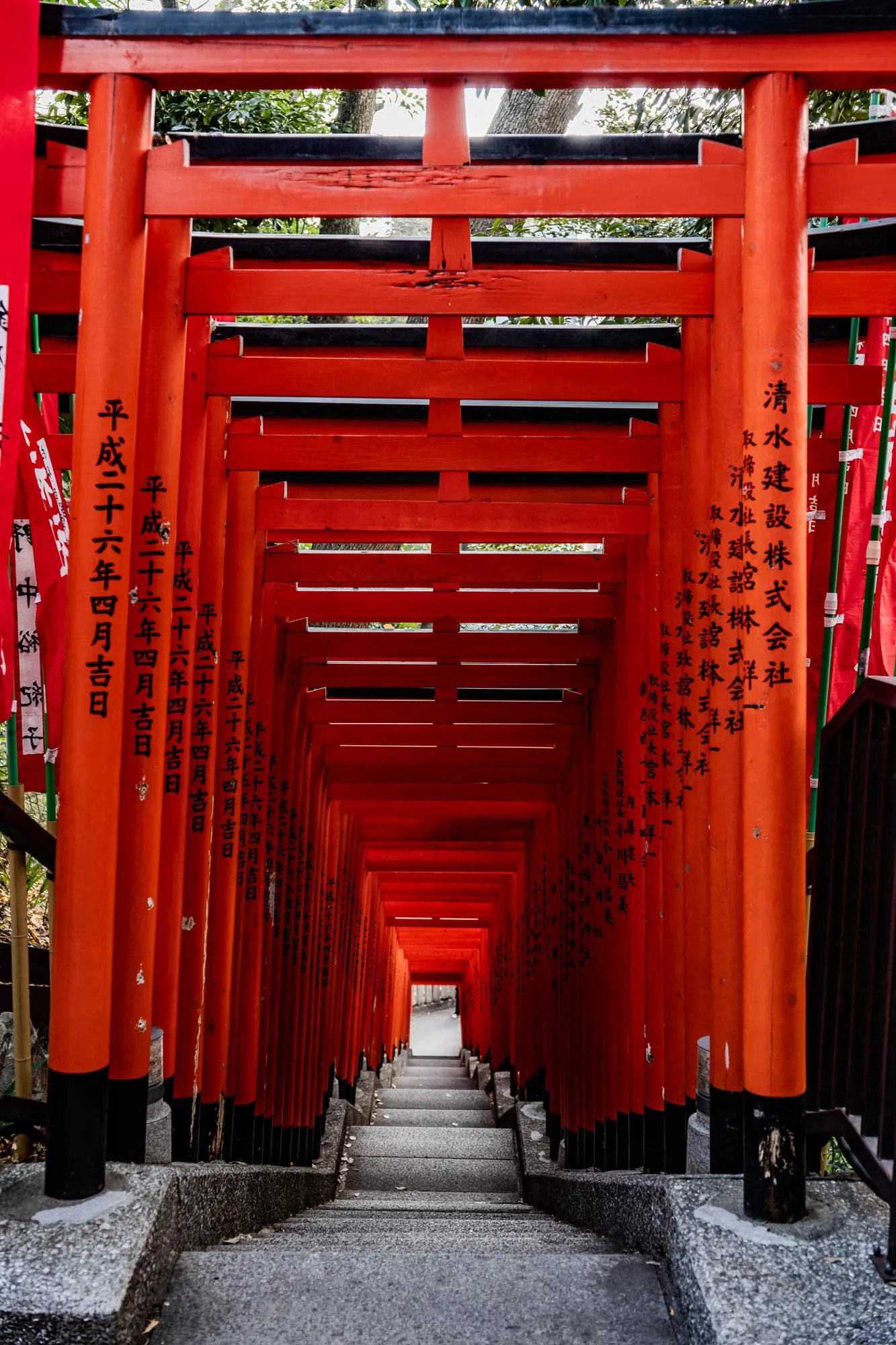 The image shows a narrow pathway lined with a series of traditional Japanese red gates called torii. These gates are arranged in a slightly curved line, creating a tunnel-like effect. They are all the same size and shape, with a black horizontal beam near the top and a black horizontal beam near the bottom. Each gate is attached to the next by a small section of grey concrete. The gates are painted red and black, with a slightly textured wood finish. The gates are stacked up on top of each other, creating an upward path. The path is lined with grey concrete slabs and some small, fallen leaves. It's impossible to see the end of the path, but it looks like it continues straight up.  The gates have writing on them, but it's hard to read. The writing is in black Japanese characters. 
