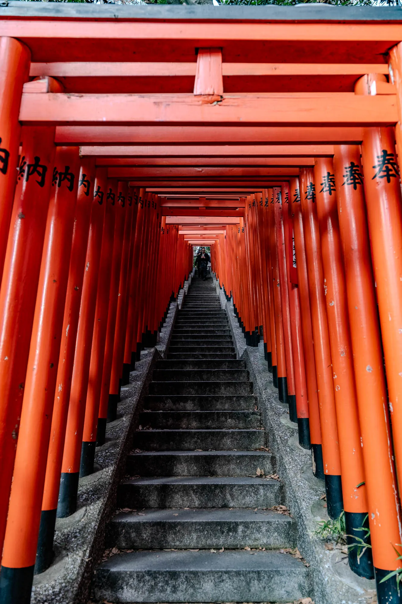 The image shows a long, narrow pathway lined on either side by traditional Japanese red torii gates. The gates are stacked on top of each other, creating a tunnel-like effect.  The path is made of concrete steps and leads upwards. The steps are grey and uneven, with some leaves and debris scattered on them. The torii gates are painted a bright orange, with black accents at the bottom. They are lined up in a perfectly straight line, creating a sense of symmetry. There are black characters written on the gates, but it is difficult to make out what they say. In the distance, there is a faint glimpse of a person walking up the stairs. The overall image is one of tranquility and serenity, with a sense of mystery and wonder about what lies beyond the gates.