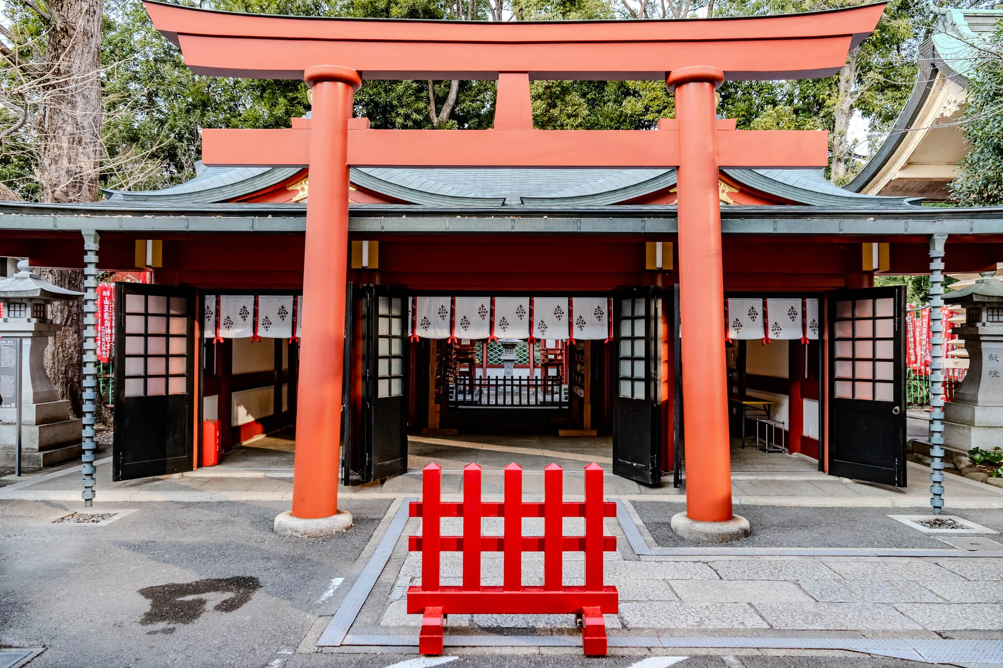 The image shows a traditional Japanese shrine with a red torii gate, a small red fence, and a paved path leading to the entrance. The torii gate is tall and wide, with two thick, red pillars that support a horizontal beam. The gate is painted a vibrant red, and the horizontal beam has a slightly curved shape. The entrance to the shrine is flanked by two black wooden doors, which are partially open. The doors are made of multiple panels, with a smaller window-like section at the top. The doors are partially covered by white fabric curtains with intricate designs. Behind the entrance, there are more red and black structures that seem to be part of the shrine complex. The path leading to the entrance is paved with gray stones, and there are a few small trees and shrubs growing around the edge of the path. In front of the entrance, there is a small red fence, also made of multiple panels. This fence is made of a very bright, almost cherry-red color and has a simple, almost utilitarian design. It is positioned on the path, creating a barrier but not blocking access. This image captures the traditional architecture of a Japanese shrine, with its vibrant colors and intricate details, creating a sense of peace and tranquility.