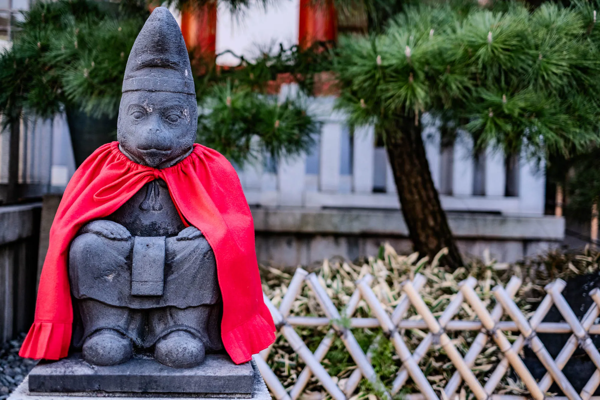 The image shows a grey stone statue of a monkey wearing a red cape and a grey conical hat. It’s seated with its arms folded. The statue sits on a stone base. In the background is a green bush with a wooden fence behind it. The fence is made of crisscrossed wooden slats that are light brown in color.  The monkey's face is visible and has a serious expression. The red cape drapes down the statue’s back and around the sides. The statue appears to be outdoors in a natural setting.  