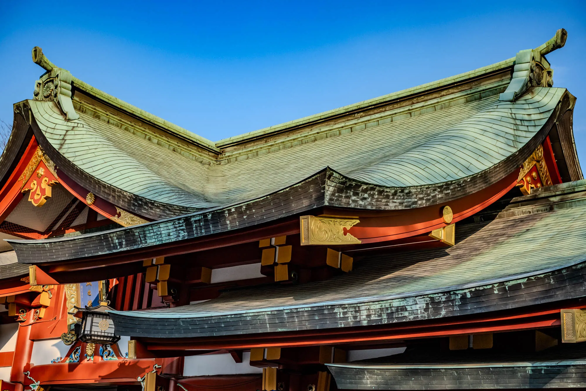 The image is a close up of the roofline of a Japanese building with a bright blue sky in the background. The roof is made of a green and black tiled material and is very intricate and curved. It features red painted beams, gold trim, and a dark wooden structure underneath the tiles. The roof appears to be very old and weathered, with evidence of wear and tear on the tiles and beams.  There is also a lantern hanging down from the underside of the roof near the bottom of the image, which is adorned with white and gold designs.  The image shows that the building is large, likely very old, and very detailed, with its intricate design and craftsmanship.