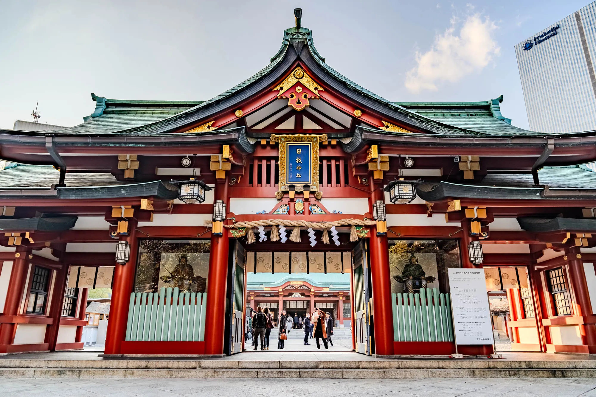 The image shows the entrance to a red and gold Japanese temple with green tile roofs. The temple is located in a city, as a tall office building with a blue sign saying "Prudential Financial" can be seen in the background. The entrance is a large gate with two red pillars on either side. The gate is adorned with ornate carvings, and there are golden accents throughout. The gate is open, and a group of people are walking through it. The temple is surrounded by a stone walkway and there are a few trees on either side of the gate. Above the gate, there is a sign that reads "日枝神社," which is likely the name of the temple.  There are lanterns hanging on either side of the entrance and a sign is to the right of the entrance containing Japanese text.   A rope with white tassels hangs over the entrance of the gate, and the gate appears to be made of wood.