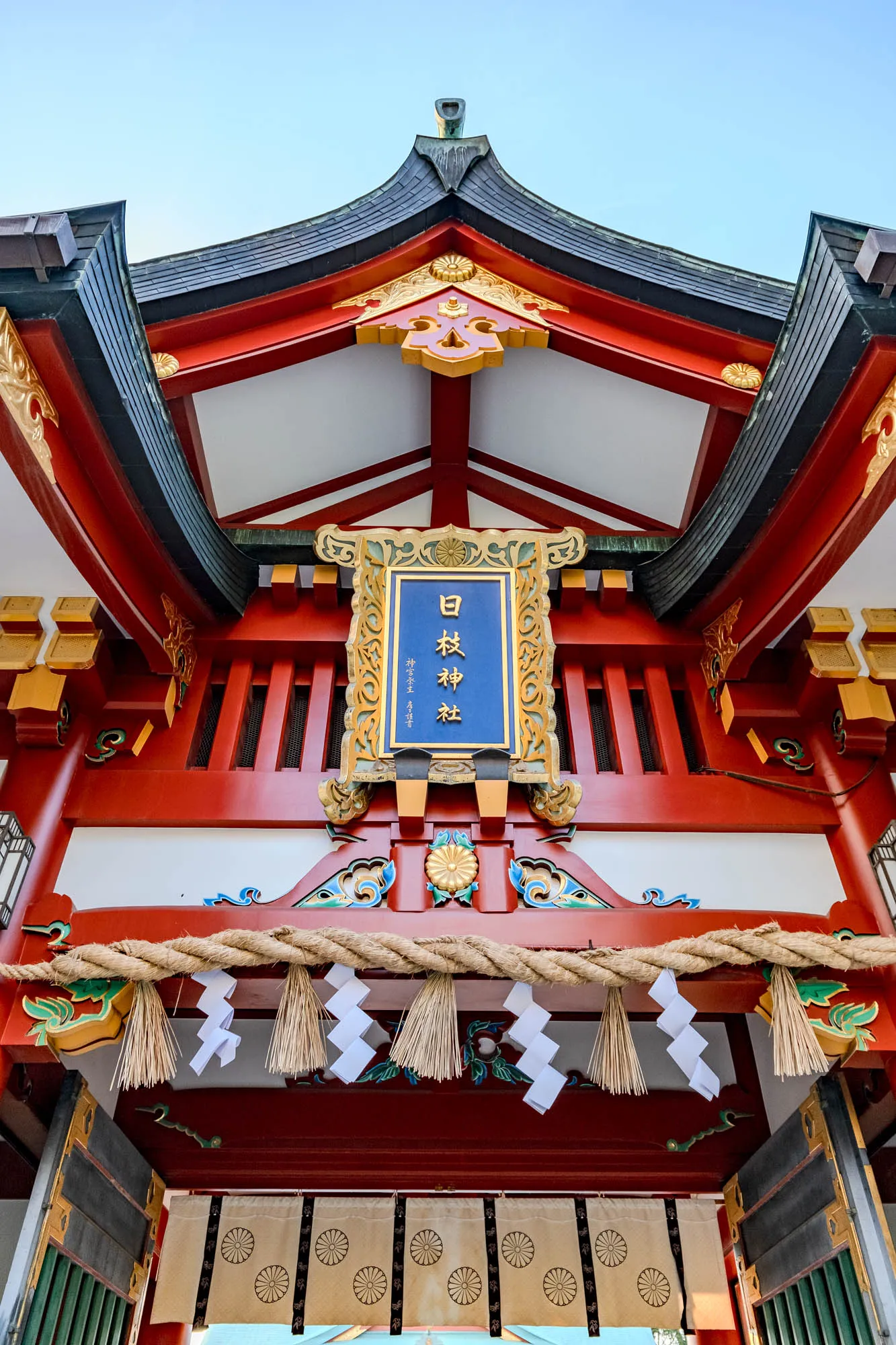 The image shows a traditional Japanese gateway, or torii, with a red and black roof and intricate gold and blue details. The torii is framed against a clear blue sky. A sign hangs from the center of the torii, featuring the words "白  神社" (Shira  Jinja) in Japanese.  The sign is framed by a carved, golden border. Underneath the sign, a thick rope hangs with white paper streamers and bundles of straw. The bottom of the torii is obscured by a large, tan curtain with a repeating motif of black circles.  The torii appears to be the entrance to a shrine or temple, surrounded by green foliage.  The scene suggests peacefulness and spirituality.