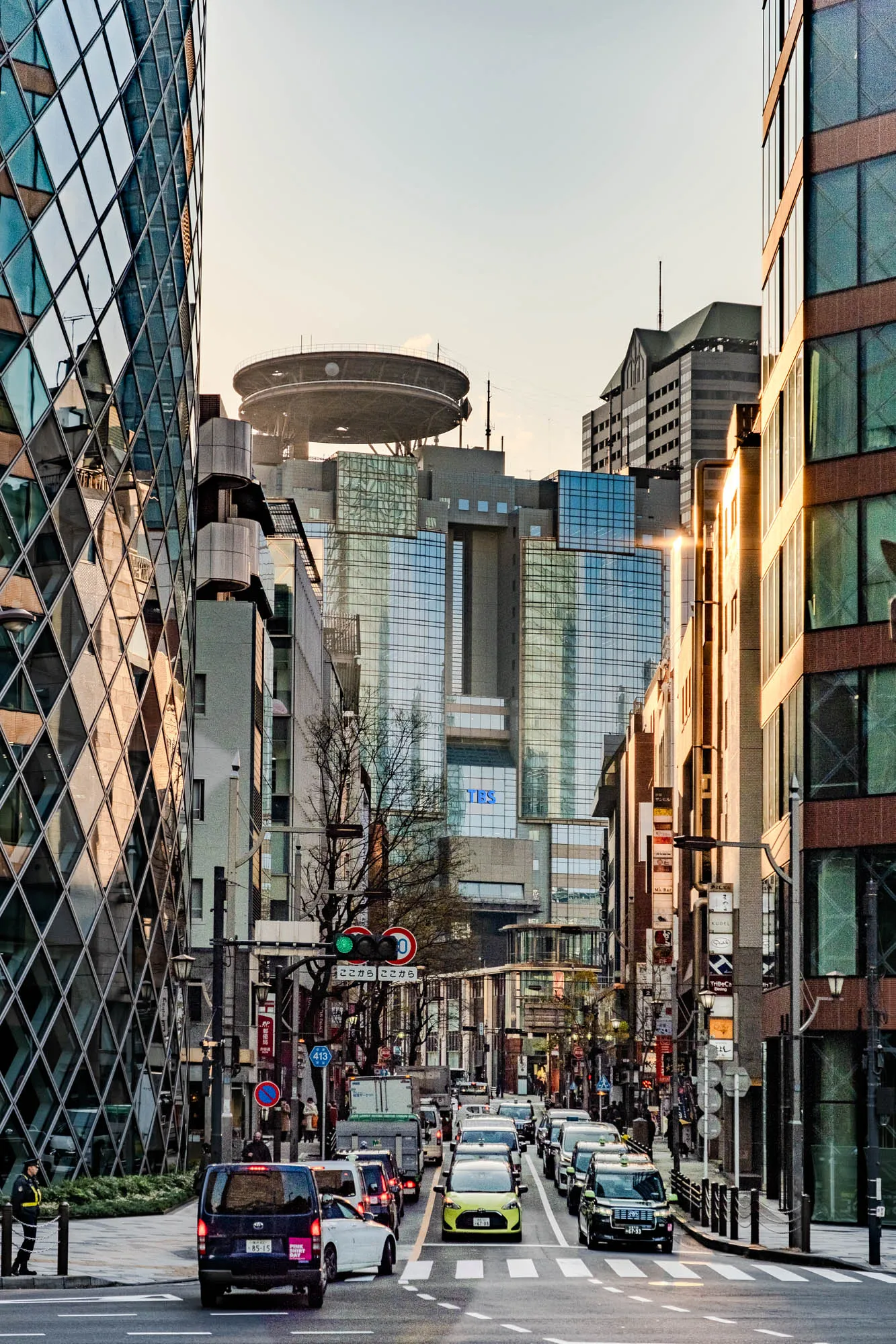 This is a photo of a city street in Tokyo, Japan. The street is lined with tall buildings, some with glass facades and some with brick. The buildings are all closely packed together, and the street is narrow and crowded with people and vehicles. The sun is setting in the background, casting a warm glow over the city. There are many cars and vans parked on the street, and several are driving down the road.  There are several pedestrians on the sidewalk, including one security guard in a uniform.  There is a large glass building on the left side of the photo with a unique geometric pattern. There is a tall building on the right side of the photo that is mostly brick.  There are several signs on the buildings, mostly in Japanese.  The street is a busy scene of urban life.