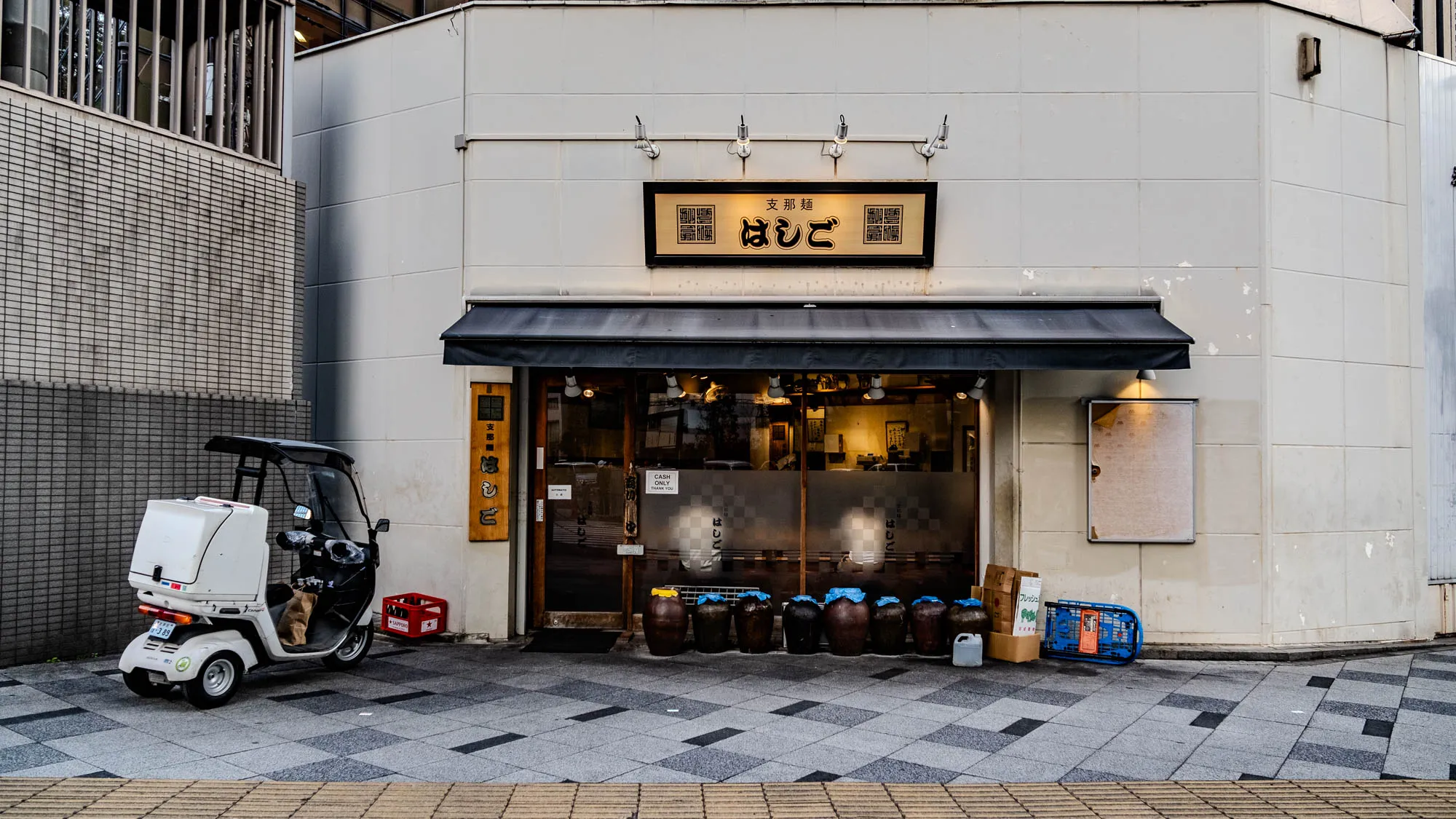 The image shows a storefront in Japan. The building is white with a black awning over the entrance. There is a sign above the entrance with Japanese characters that say "天ぷら" (tempura) and "ねぎと" (with green onions). The storefront has a large glass window that looks into the restaurant. There are a few tables and chairs visible through the window. The door is closed and there is a sign on the door that reads "CASH ONLY." There are several large brown jars lined up in front of the storefront. On the left side of the image, there is a white delivery scooter parked on the sidewalk. The scooter has a box on the back for carrying deliveries.  There are boxes of various sizes on the sidewalk as well. The sidewalk is made of large square tiles.  The scene is lit with the warm glow of afternoon sunlight. 
