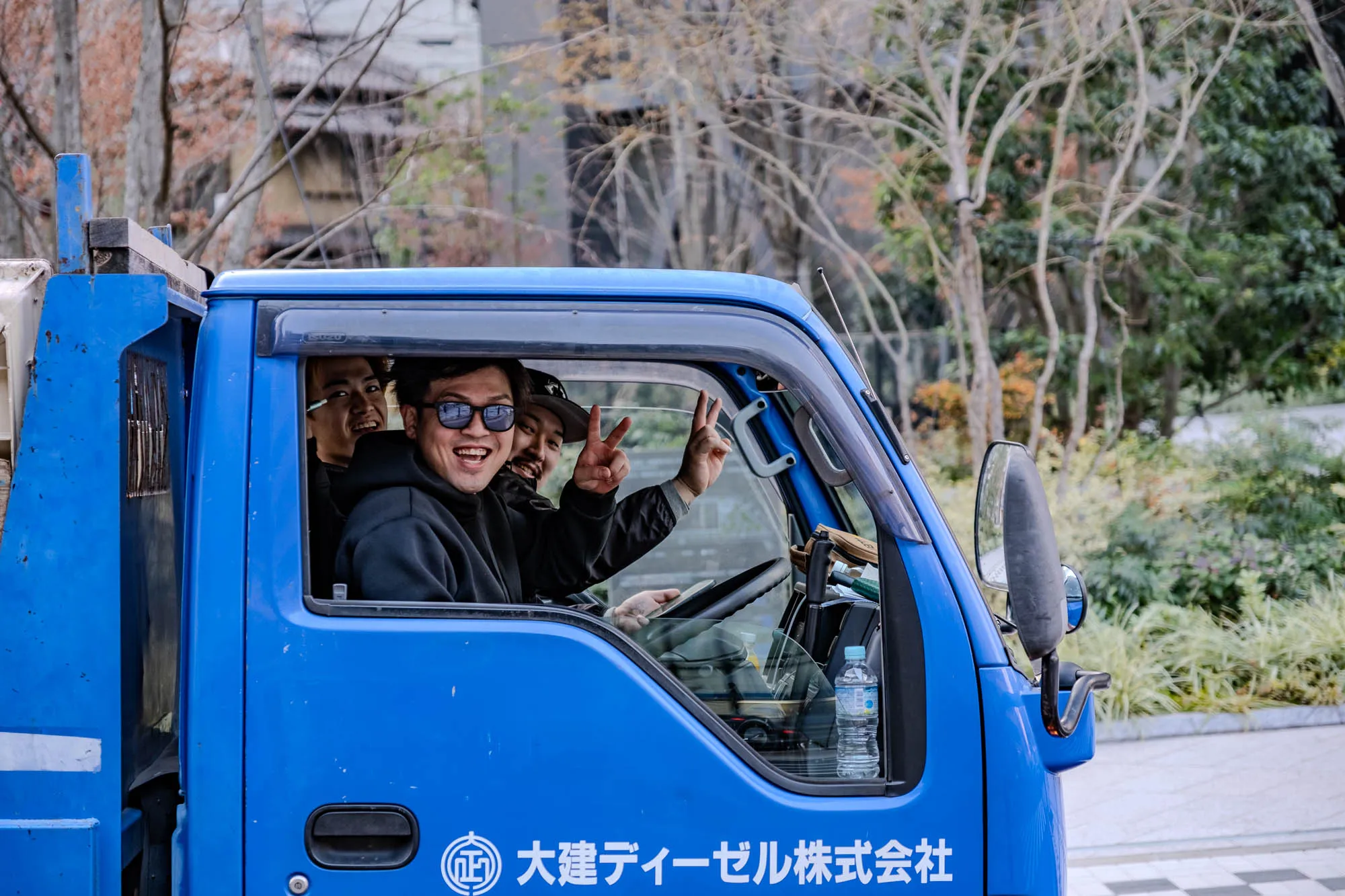 The image shows the side of a blue delivery truck. Two men are sitting in the cab. The man in the driver’s seat is wearing sunglasses and a black hooded jacket, and he’s holding up a peace sign. The other man is looking toward the camera and smiling. The truck is parked in front of a building with a green lawn behind it. The side of the truck has the words “Daiken Diesel Co., Ltd.” in Japanese and a small logo.