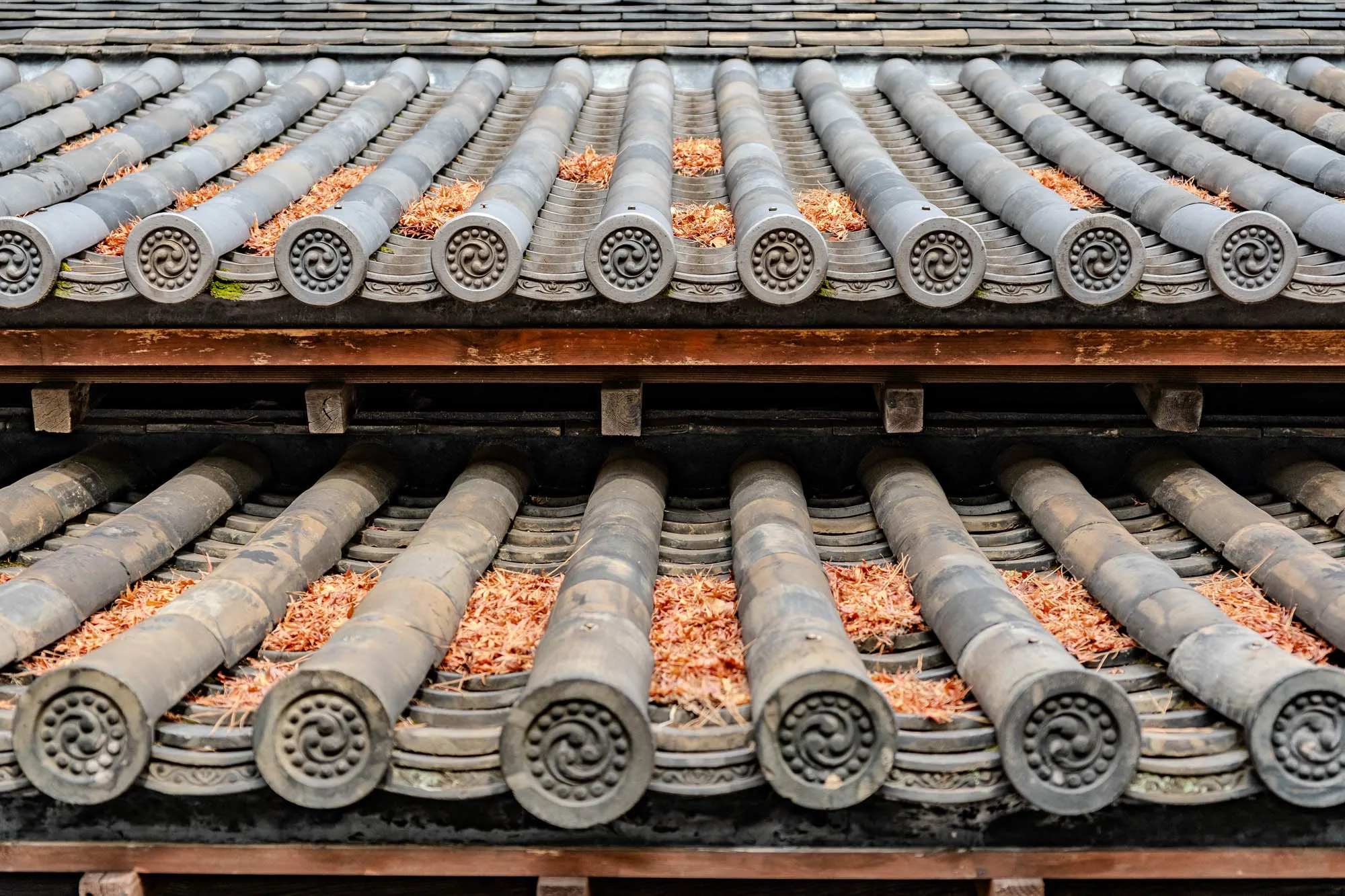 The image shows a close-up view of a traditional Japanese roof. The roof is made of dark grey tiles that are arranged in rows, each row overlapping the one below. The tiles are cylindrical in shape and have rounded ends. The tiles are adorned with intricate patterns in the center that appear to be a swirling or circular design. There is a wooden beam that runs horizontally across the roof, separating the top row of tiles from the bottom. The wood is a light reddish brown. There are also little bits of dry leaves and pine needles that have fallen onto the roof and rest on the surface of the tiles. The overall impression is of a weathered but well-maintained roof that gives a sense of age and tradition.