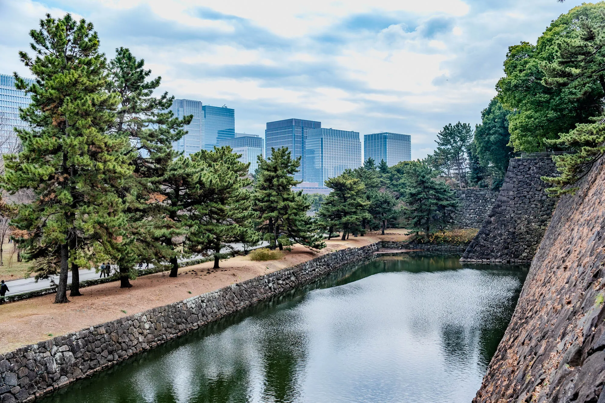 The image shows a view of a moat in front of a stone wall. The moat is filled with still water, and the wall is made of large, irregularly shaped stones. The wall is quite high, and a few people can be seen walking along the path at the top of the wall. There are several trees growing along the edge of the moat, and more trees are visible in the distance. Behind the trees are several modern skyscrapers. The sky is cloudy and overcast.