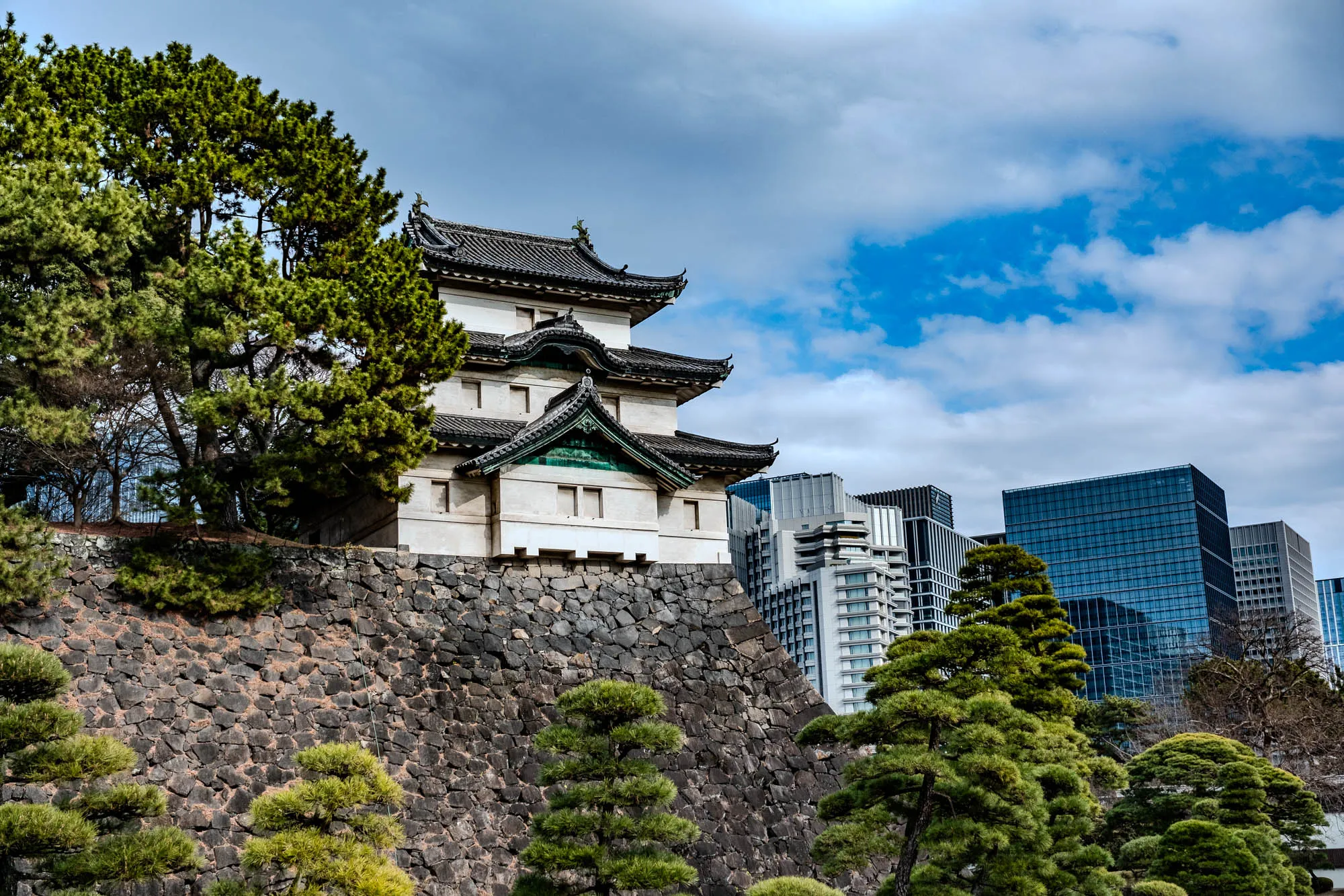 The image shows a Japanese castle building with a black tiled roof.  It has a white exterior with green trim on the eaves and a few windows. The building sits atop a high stone wall that is made of irregularly shaped stones.  Behind the building, several modern office buildings are visible, and a few tall trees are in the foreground. There is a bright blue sky with white clouds.  The overall effect is a contrast between traditional Japanese architecture and the modern cityscape.