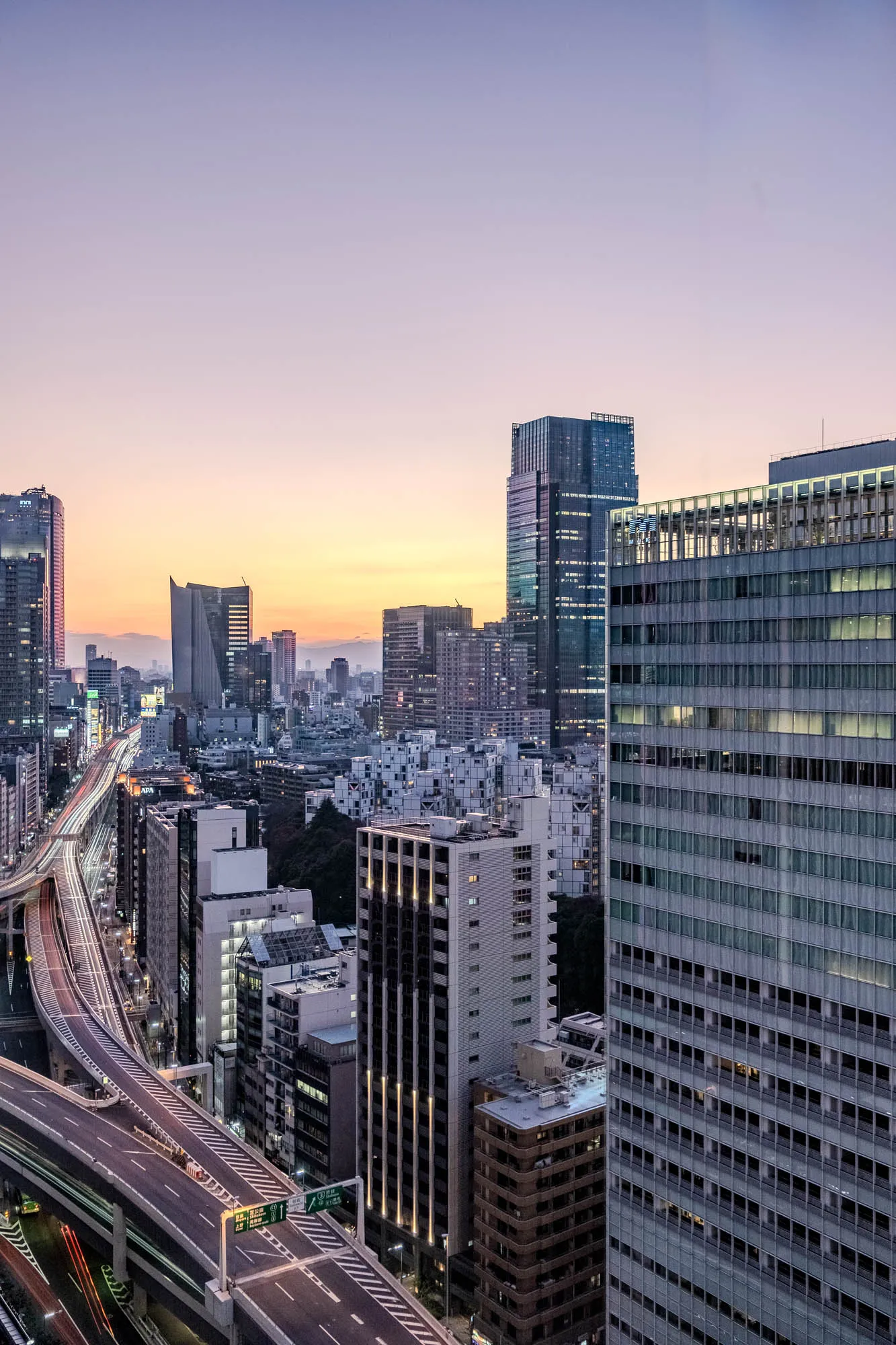 This image is a high-angle view of a city skyline at dusk. The image is dominated by tall buildings. In the foreground, there is a highway with multiple lanes of traffic. The traffic lights streak across the highway as cars travel along the road. You can see the buildings on either side of the road. The buildings in the background are illuminated with lights. The sky is a soft purple color, indicating the end of the day.