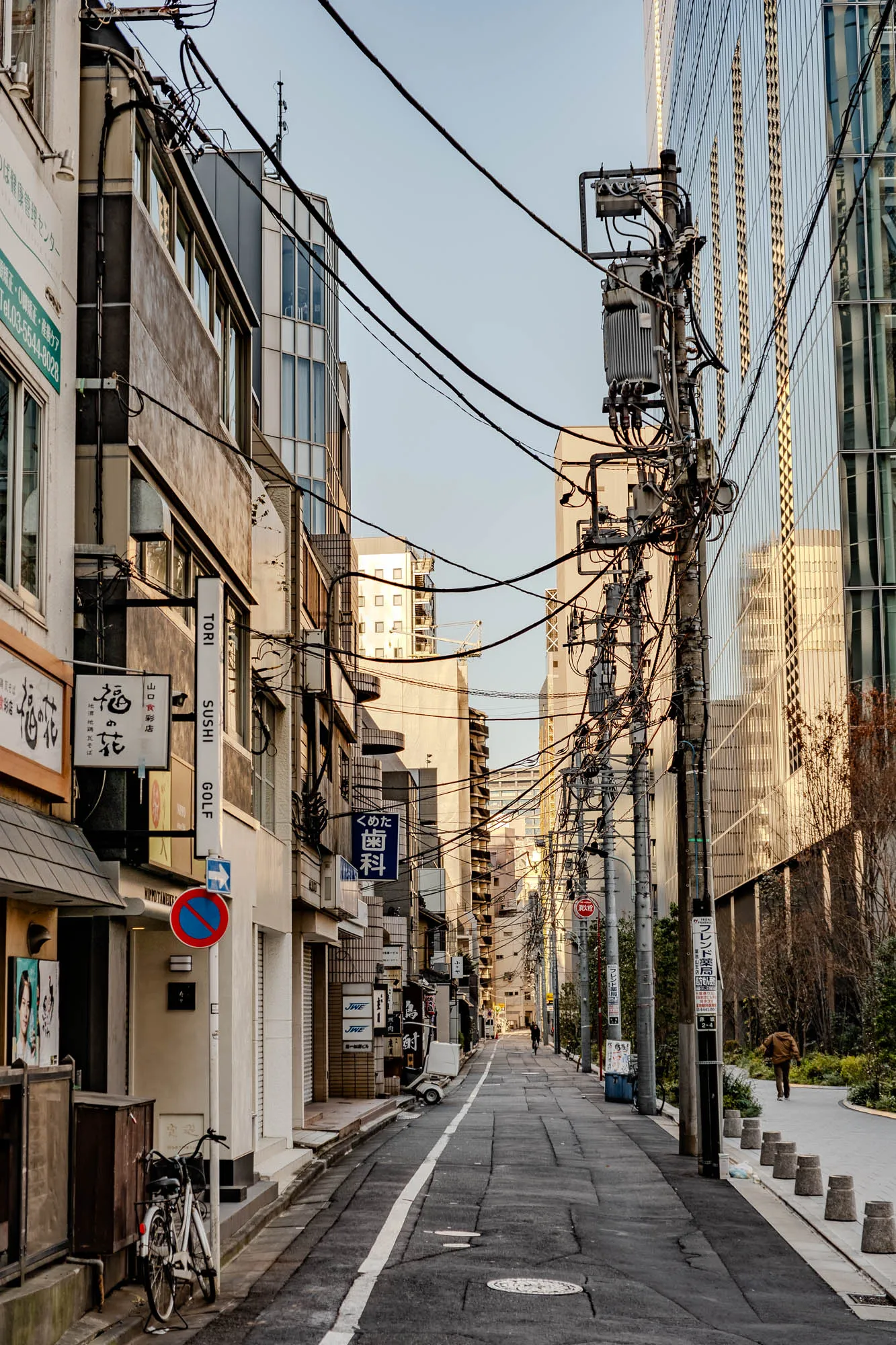 This is a photograph of a narrow street in a city, likely in Japan. The street is lined with buildings on both sides, with power lines strung overhead. The buildings are a mix of traditional and modern styles, with some having storefronts and signs in Japanese. There are a few signs in English, including "Tori Sushi Golf". The buildings are made of concrete and brick and have a weathered appearance. The street is paved with asphalt and has a white line down the center. The sky is overcast and the street is empty except for a single bicycle parked near the edge of the sidewalk. There is a trash can in the street, and there are some plants growing along the edge of the sidewalk. The buildings in the background are tall and have a modern glass facade. On the right side of the image, the edge of the sidewalk extends into a park with a small patch of trees.