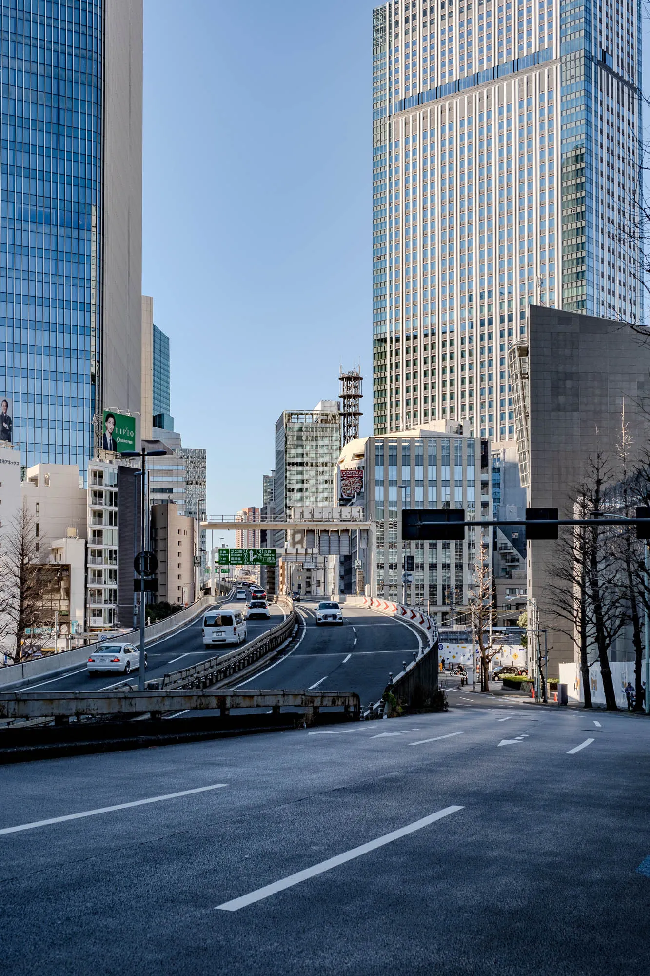 The image shows a highway in a city with tall buildings on either side. The highway curves to the left in the foreground and continues in the distance. There are a few white cars driving on the highway. In the background, a large skyscraper with many windows stands tall. To the left of it, there is a smaller building with a billboard on the side. The sky is a clear blue and the image has a bright and sunny feel. 
