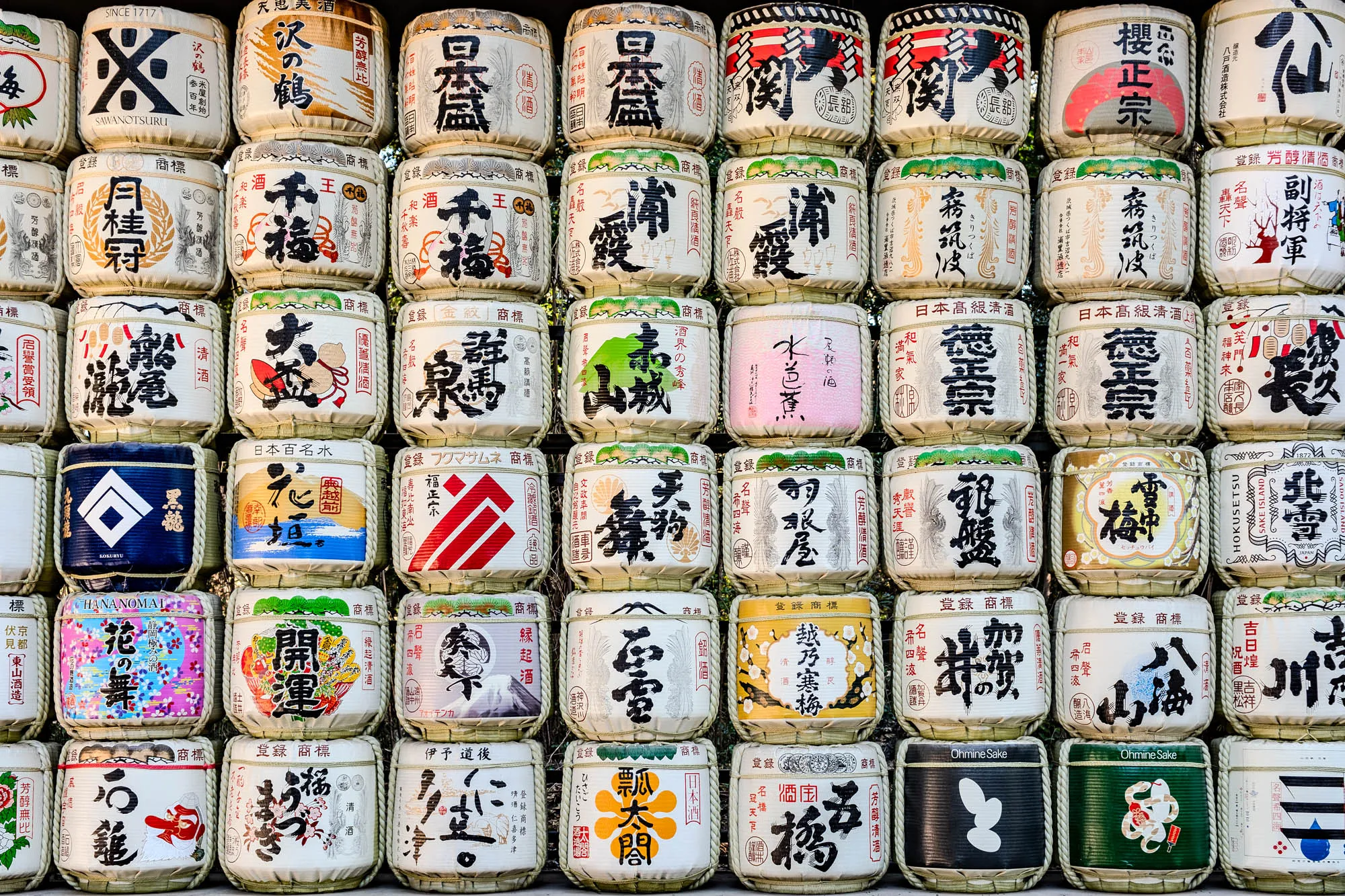 The image is of a wall covered in rows of sake barrels.  The barrels are all made of wood and have a straw covering.  Each barrel has a unique label with Japanese text and images. The labels depict things such as flowers, mountains, sake bottles, and traditional Japanese designs. Some of the text on the labels include the words "sake," "Japanese," and "since 1717."  There are 4 rows of barrels with 12 barrels in each row, for a total of 48 barrels.