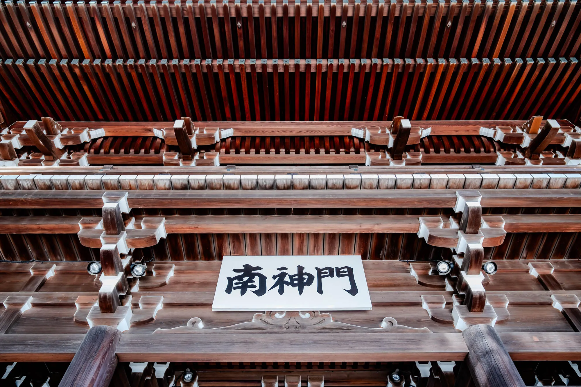 The image shows the underside of a traditional Japanese wooden structure. It appears to be a temple or shrine, with intricate details and a sign with Japanese characters. The ceiling is made of many horizontal wooden planks, which run parallel to each other. Above the planks are more wooden beams. The sign has black characters on a white background.  There is a decorative wooden motif below the sign, with white painted accents.  There is a light fixture on each side of the sign, and a pillar on the left and right side of the image. 
