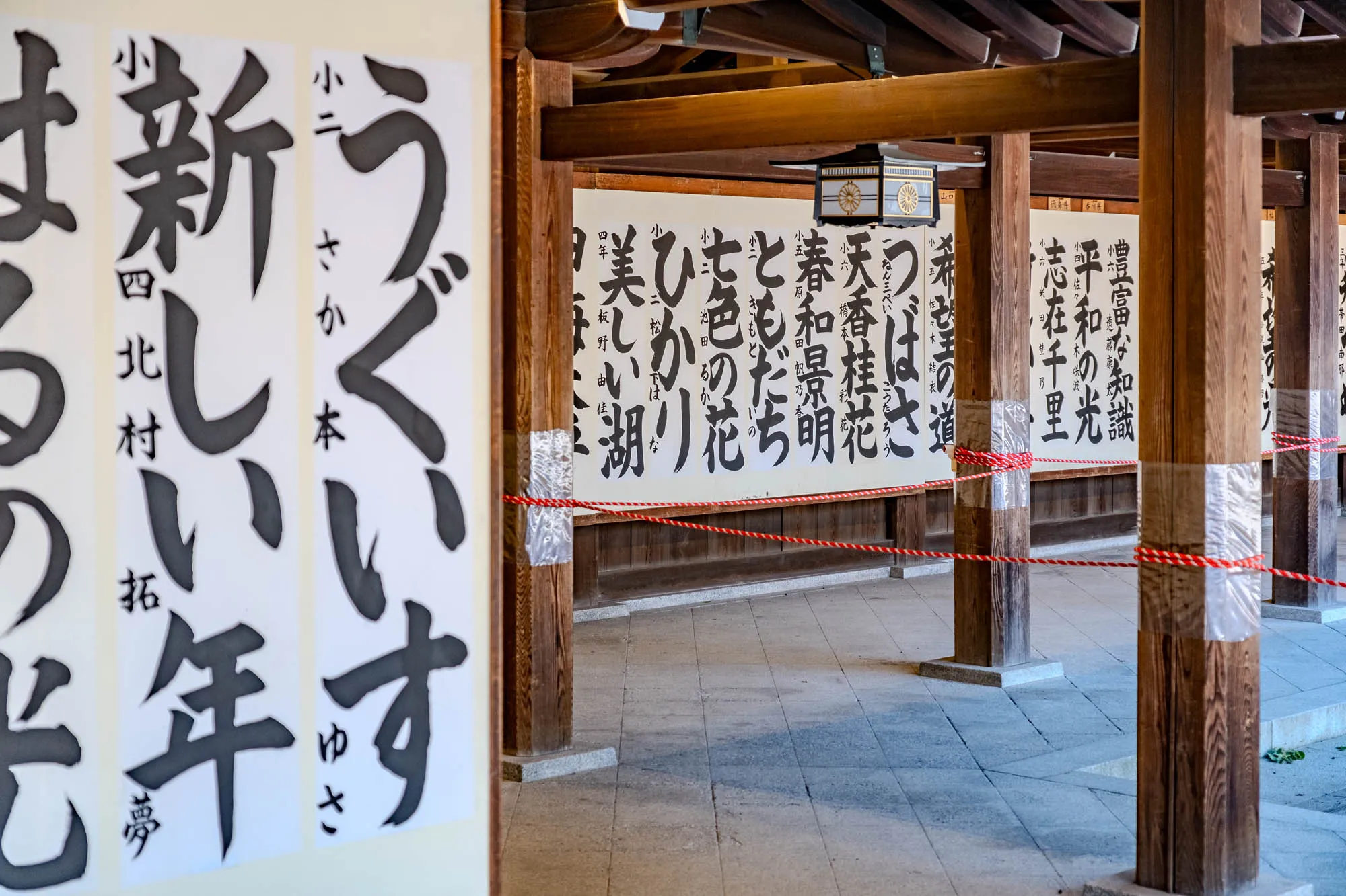 The image shows a large tree with thick branches and leaves. It has a woven rope wrapped around its trunk, and there are small white flags hanging from the rope. In the background, there is a wooden fence with many small wooden plaques hanging from it on orange strings. The plaques are all different shapes and sizes and have writing on them in a language that cannot be read in this image. The fence is made of horizontal wooden slats and the area behind it appears to be dirt and grass.