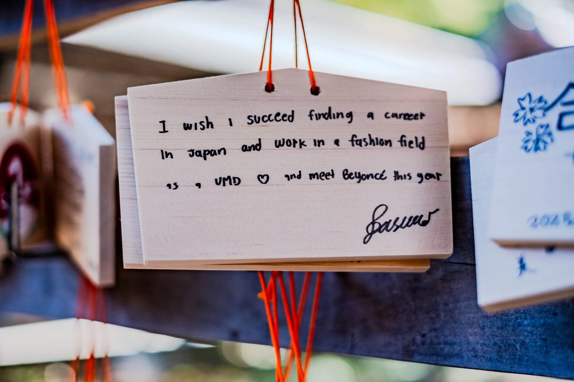 The image shows a small wooden plaque hanging from orange string. The plaque is made of light wood with a grainy texture. It is rectangular and has rounded edges at the top. On the plaque is black ink writing that reads "I wish I succeed finding a career in Japan and work in a fashion field as a UMD ♥ and meet Beyoncé this year" with a signature at the bottom that reads "Sasuns". There are other plaques behind it hanging from the same type of string, the one directly behind it also has black writing but is out of focus.  There is a partially visible plaque to the right of the main plaque that has a drawing of a flower in blue ink on it. The background is blurry and out of focus.