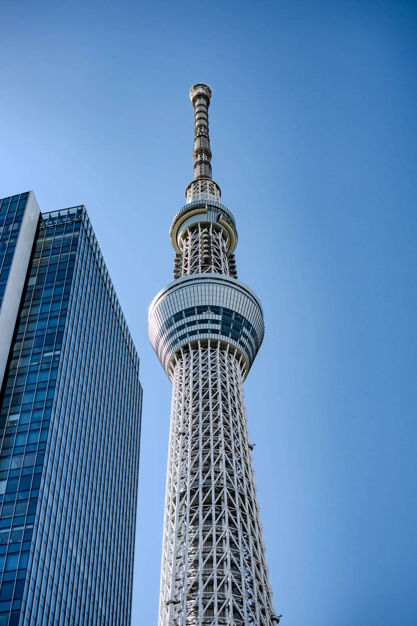 The image shows a tall, white tower against a clear blue sky. The tower is made of steel, with a lattice-work design and a large, round platform at the top. The top platform has a white railing with small, dark accents.  There is a tall, thin rectangular building on the left side of the tower. It is mostly blue, and the windows are dark. The building and tower stand out against the bright blue sky.  