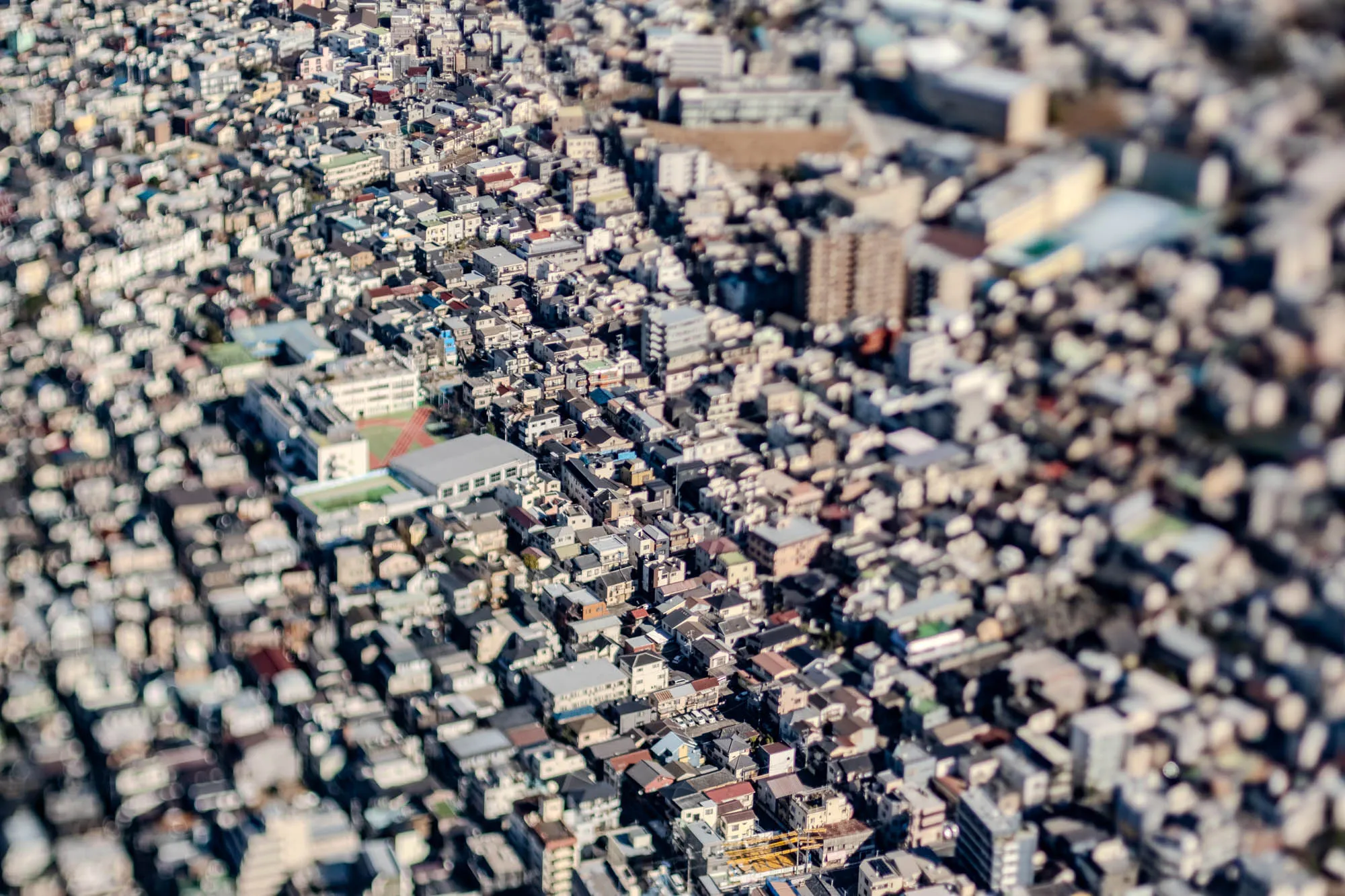 This image is a blurry, aerial view of a densely packed city. There are many buildings of various sizes and shapes, most of them are either grey or white. In the middle of the image, there is a large building with a flat, green roof. The buildings are mostly out of focus, making it appear as if the city is a miniature model. There are also some trees scattered throughout the city, but they are also out of focus.