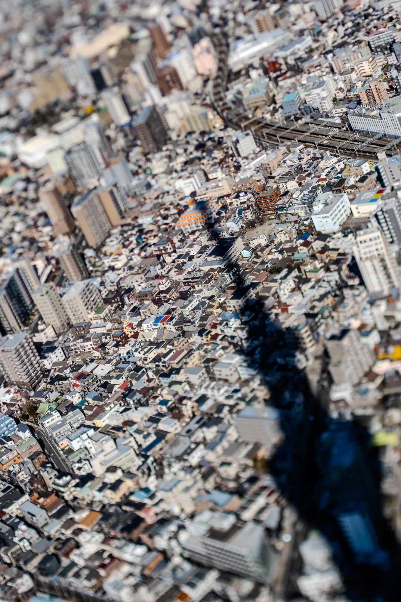 An aerial view of a densely packed cityscape.  The image is taken from a high angle looking down.  The buildings are small and clustered tightly together, giving the impression of a model city.  The buildings are primarily residential and are mostly grey and brown with a few pops of brighter color.  A large, dark shadow from a tall building casts over the smaller buildings in the image.  A train track, with a train running on it, can be seen in the distance.