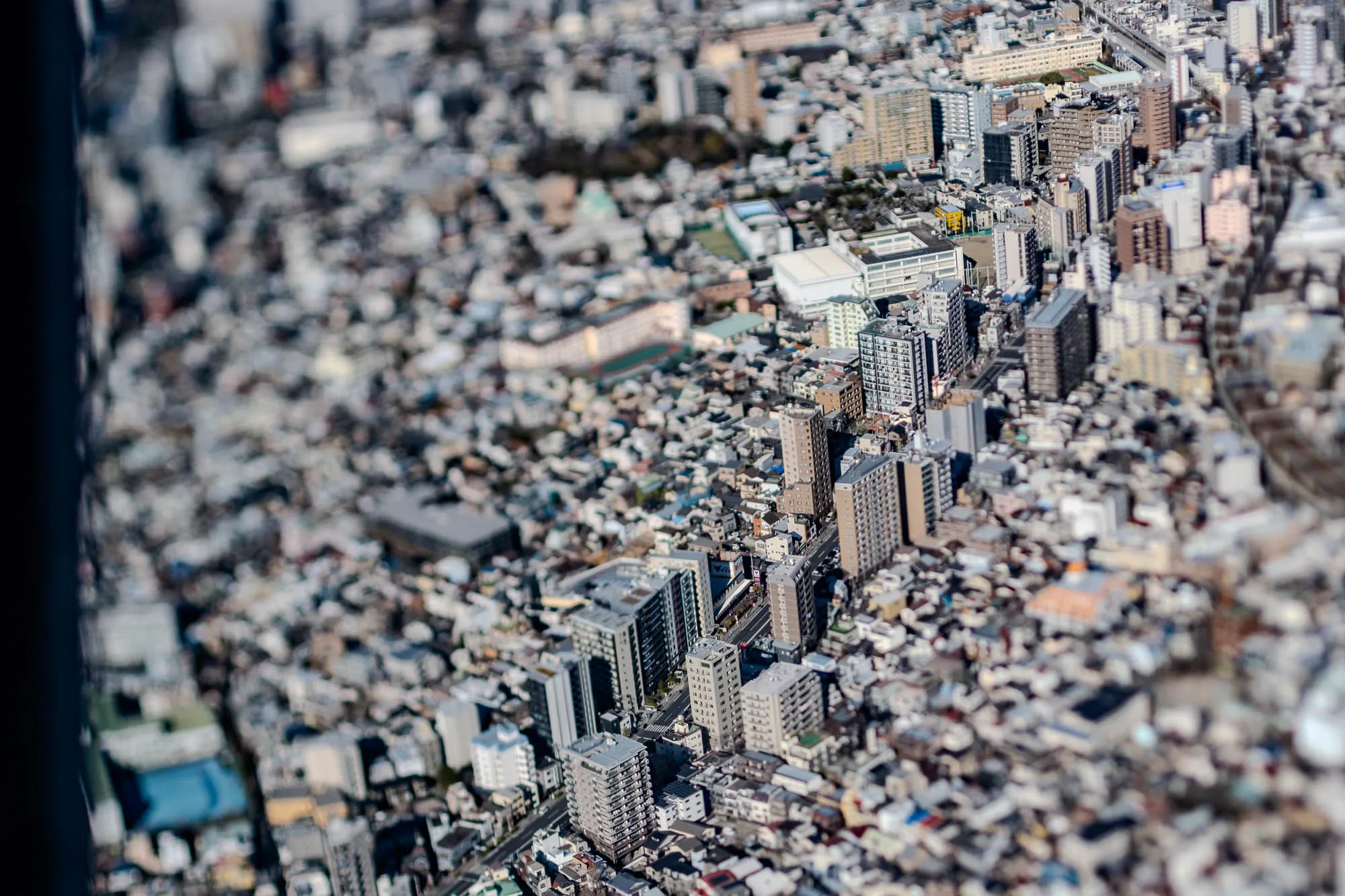 A blurry aerial view of a city with tightly packed buildings of various heights. The view is taken from a very high vantage point, giving the impression of a miniature model city. The buildings are mostly a light grey or tan color, and there are patches of green vegetation scattered throughout the city. The photo is slightly out of focus, adding to the miniature effect. There is a black vertical bar in the left side of the image. The perspective is from above the city.