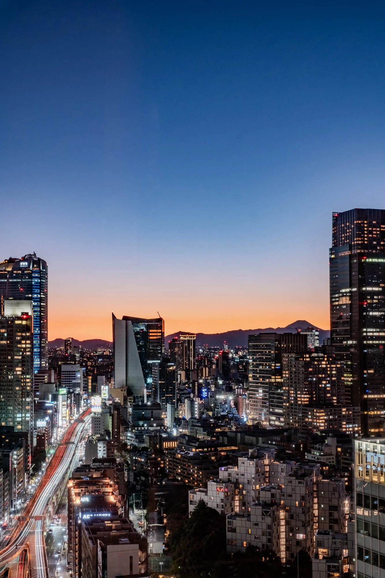 The image shows a nighttime cityscape with tall buildings and a highway. The sky is a dark blue with hints of orange from the setting sun. In the foreground, a highway with streetlights and speeding cars stretches out into the distance. To the right of the highway, a cluster of buildings with illuminated windows stand in the foreground. Behind these buildings are many other structures, all lit up.  A tall skyscraper dominates the right side of the image, with the top illuminated. Behind the cityscape, a dark mountain range stands out against the colorful sky. 
