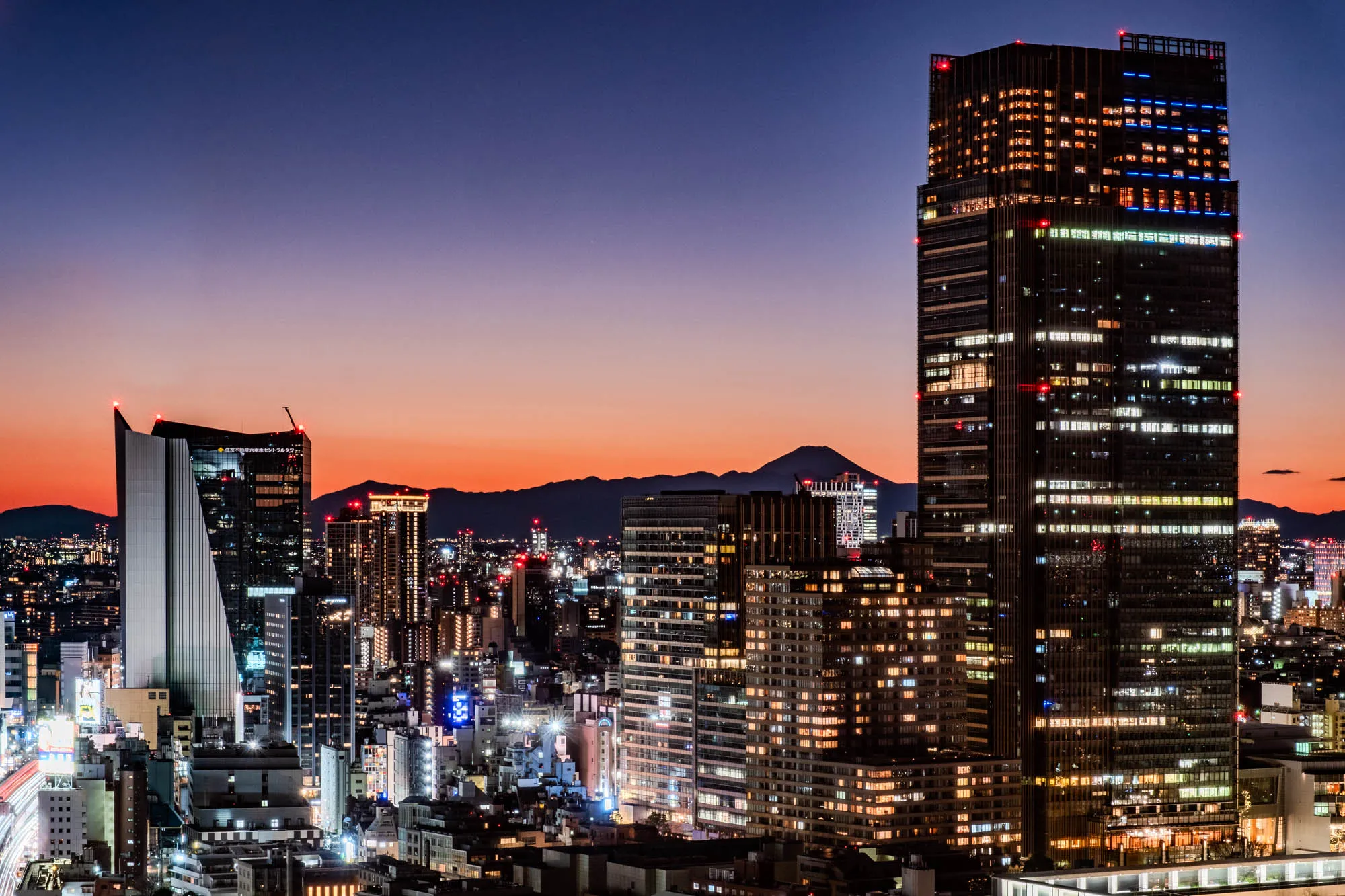 A high angle view of a city skyline at night. In the foreground is a busy highway, with a long exposure showing streaks of light from cars. On either side of the highway are tall buildings with many windows. The sky is a deep blue, with a hint of orange on the horizon. In the distance, a mountain range is silhouetted against the sky. The city is lit up with many lights, creating a beautiful and bustling atmosphere.