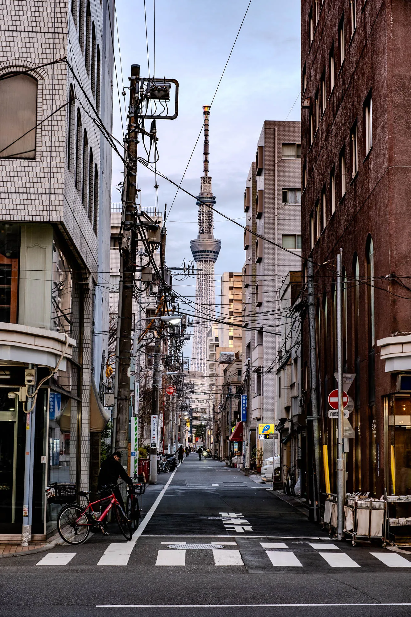 This is a photo of a narrow street in a city. There are buildings on both sides of the street and a tall tower in the distance. The street is paved with black asphalt and there are white painted lines indicating a crosswalk. There are many power lines overhead and there is a metal pole with wires running across the street. On the left side of the street there is a man standing next to a red bicycle on the sidewalk. The building on the left is a multi-story brick building with a storefront and windows. The building on the right is also brick with a storefront but is less tall. The tower in the distance is a large, slender structure that tapers at the top and is made of a metal structure. It appears to be a radio or television broadcast tower. There are several smaller buildings in the background between the street and the tower. The sky is clear and slightly overcast. The photo was taken during the day. 
