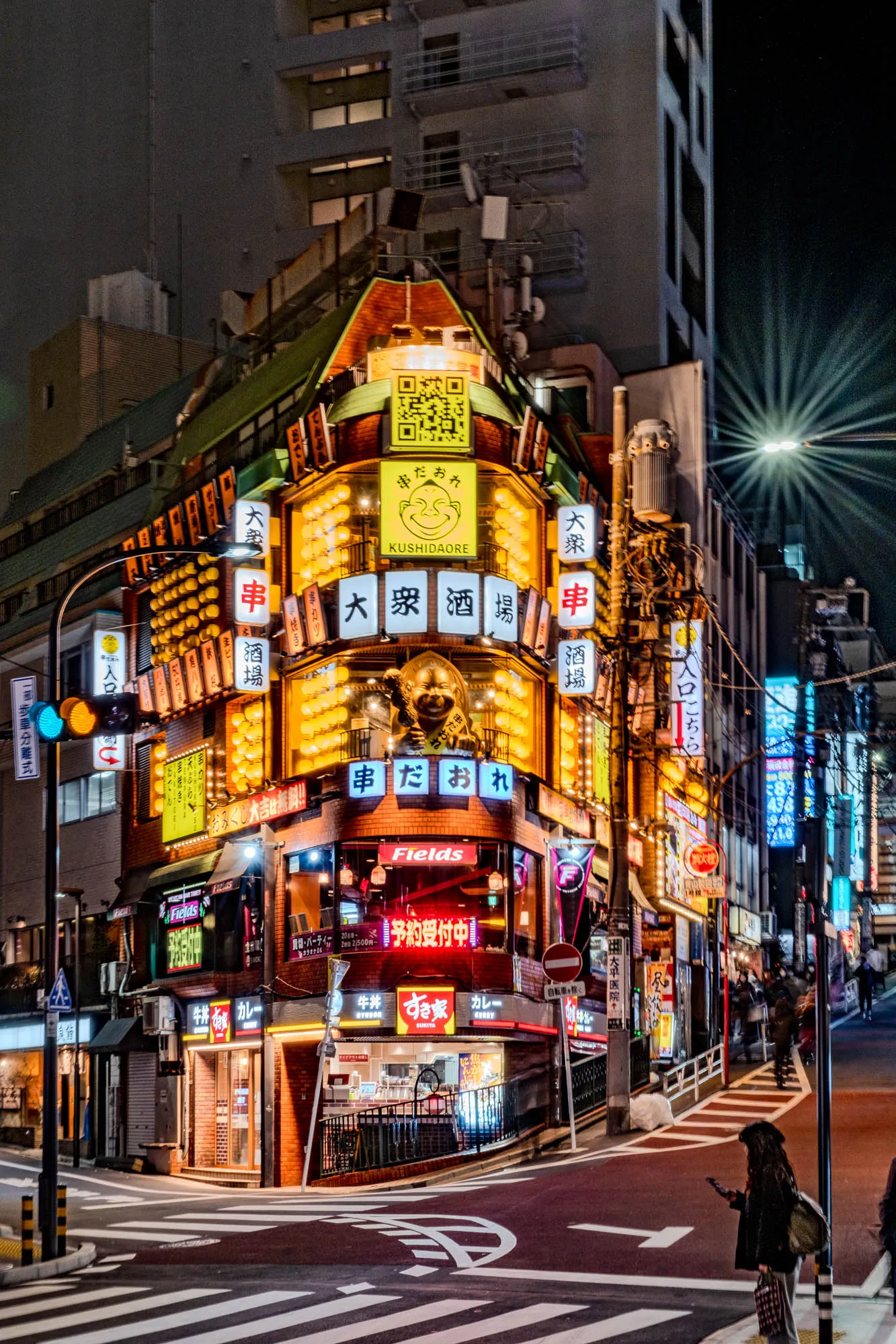 The image is a nighttime street scene in Japan. There is a corner building with a variety of neon signs in Japanese, along with several other buildings in the background. The building on the corner has a variety of signs on it, including the words "KUSHIDAORE", "Fields", "大衆串酒場" (which translates to "Popular Kushiyaki Bar"), and  "牛丼すき家 カレー" (which translates to "Gyudon Sukiya Curry"). There is a crosswalk in the foreground with a person walking across it. The street is lit up with streetlights and the signs on the buildings.  The building on the corner also has a large metal statue of a smiling man wearing a hat with a name tag that says "まだおれ" (which translates to "Still Here"). The ground in the foreground is paved with a cobblestone pattern. The street looks bustling and vibrant.  