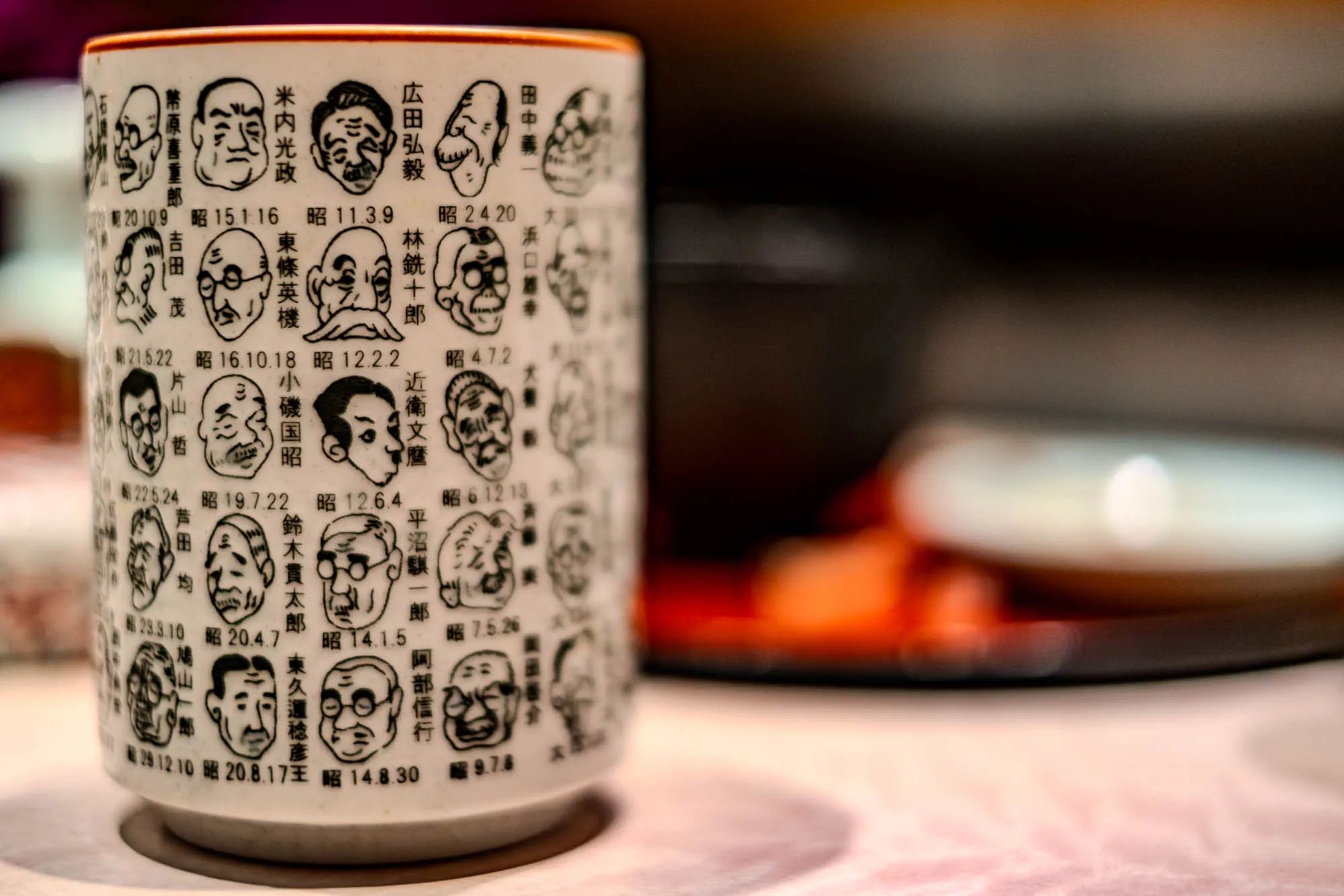 The image is of a white ceramic cup with a brown rim. The cup is covered in black and white drawings of men's faces, each with a number and a date below. The cup is sitting on a table, and there are some blurry out of focus objects in the background, including a white saucer and a blurry red object. The date written below each face is in the format 昭X.X.X where X represents a number. Some of the men are wearing glasses and some have mustaches.