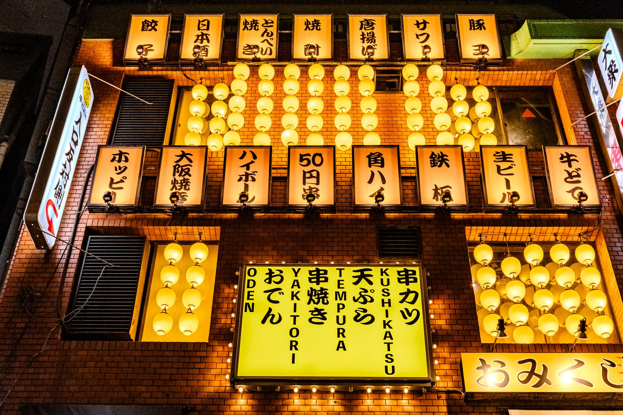 The image shows a brick building in Japan at night. There are many glowing yellow signs, hanging lights and a bright yellow sign in the center. The signs on the upper level appear to be advertising food. The signs on the lower level are for various Japanese foods including, "串カツ" (kushikatsu) or skewered deep-fried food, "天ぷら" (tempura) or deep-fried seafood and vegetables, "串焼き" (kushiyaki) or skewered grilled food,  "焼ハイボール" (yaki highball) or a type of Japanese whisky drink, "50円" (50 yen) and “大阪燒" (Osaka-yaki) or a savory pancake with many ingredients. There are also signs for "日本酒" (nihonshu) or Japanese sake and  "ホッピ" (Hoppi) which is a popular Japanese beer brand. The center sign says "おでん" (oden) which is a popular Japanese winter dish of various ingredients stewed in a flavorful broth.  The sign also says "YAKITORI"  "TEMPURA"  "KUSHIKATSU"  "ODEN"  "おでん" (oden) "天ぷら" (tempura) "串焼き" (kushiyaki) “焼ハイボール” (yaki highball)  "50円" (50 yen) "角ハ" (kaku-ha), “生ビール” (namabiiru) or draft beer.  There is a sign on the lower right that says "おみくじ" (omikuji) which refers to fortune slips that people draw from a box. The building is lit up with warm yellow lights and the scene is very vibrant.