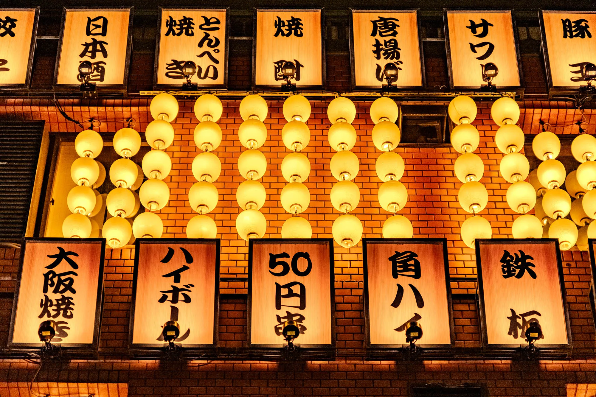 The image is a close-up shot of an illuminated brick wall with Japanese lanterns hanging from the brickwork, with signs interspersed between them, lit up with warm light, making the red bricks appear orange. The lanterns are round and translucent, giving off a soft, warm glow.  There are eight rows of lanterns across the wall, with seven in each row. The signs are all rectangular and lit up with warm white light, some have black Japanese characters on them. The signs appear to be for a Japanese restaurant. 
