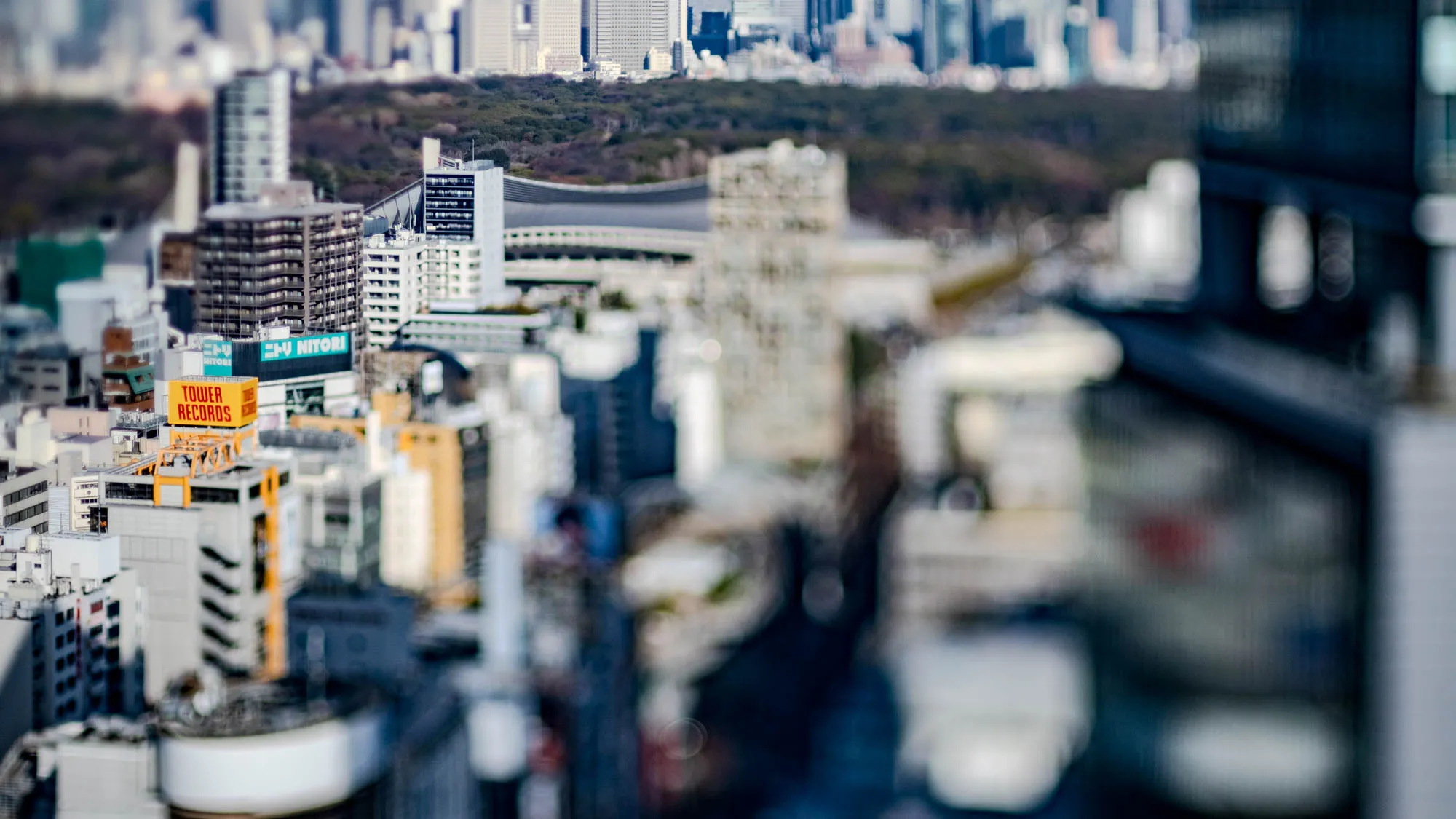 The image is a bird's eye view of a city skyline, taken from a tall building looking down on a dense urban area. The image is blurry and out of focus, giving the impression of looking through a window, or of being far away from the city. In the foreground is a large dark building, out of focus, that looks like it might be a parking garage.  In the middleground, there are dozens of buildings. One building is partially visible with a sign that says "Tower Records" in red and yellow letters.  In the background, the buildings are further out of focus and appear as a hazy, colorful, impressionistic representation of the city's skyline.