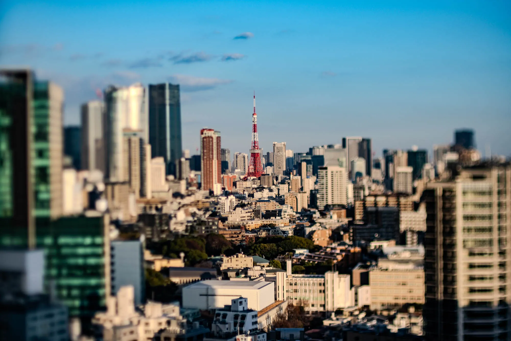 The image is a cityscape, most likely in Tokyo, Japan. It shows a wide shot of the city from a rooftop, with a famous tower, Tokyo Tower, in the background. The tower is red and white, tall, and skinny. There are numerous other buildings in the background, all blurry and indistinct. In the foreground, the viewer is looking directly at the tops of buildings, most of which are grey and concrete. The sky in the image is a clear blue, with a few wispy clouds in the distance. It's a sunny day, although the buildings cast shadows, suggesting it is early afternoon or late morning. The image is likely taken with a wide-angle lens, emphasizing the vastness of the city and the dominance of the Tokyo Tower.