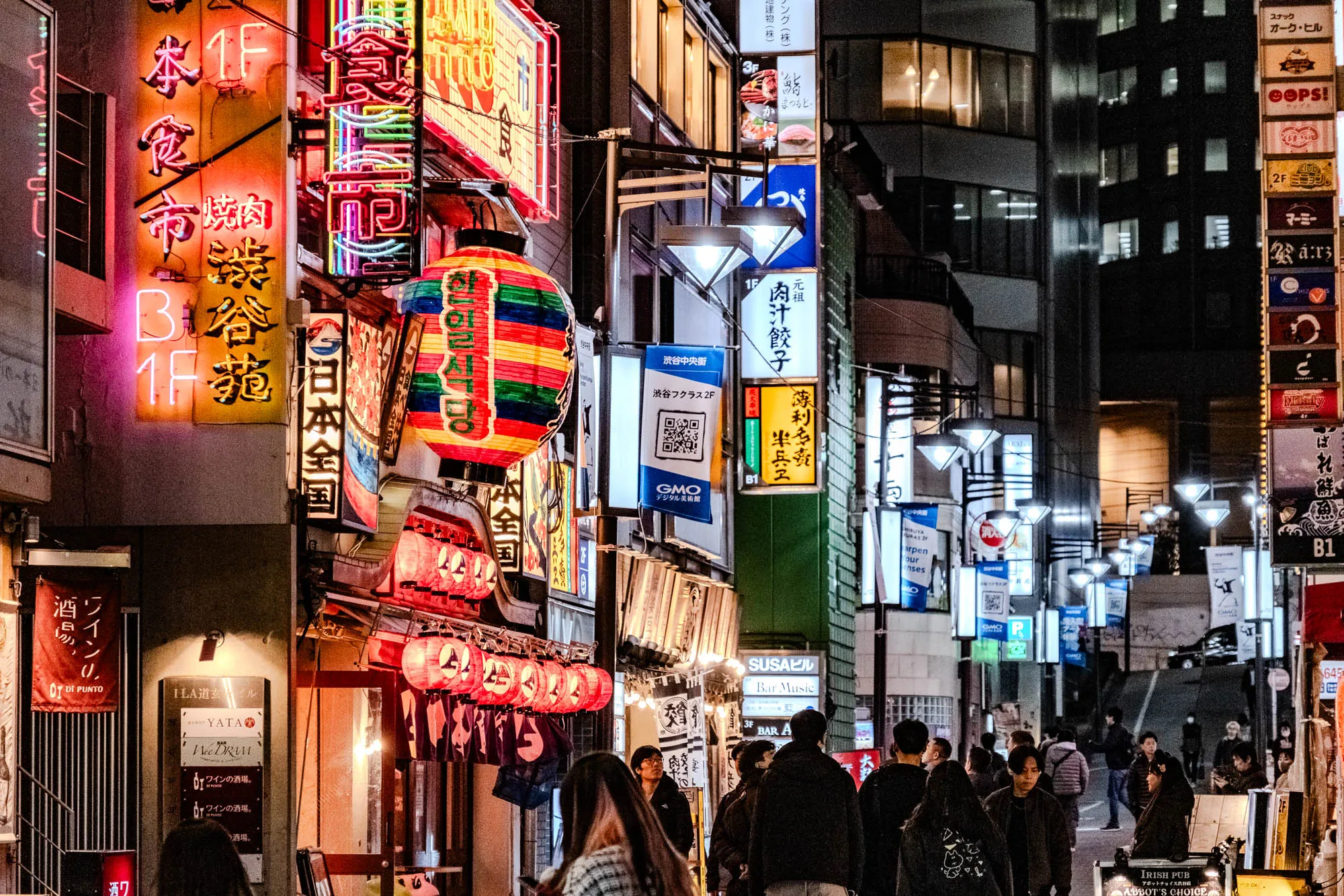 This image shows a narrow, bustling street lined with buildings in Japan. The scene is lit by bright, neon signs in Japanese and English. On the left side of the street, the building's facade is covered in large, brightly colored neon signs featuring Japanese characters that likely advertise local businesses.  The text "B 1F" is visible, likely indicating basement floor levels. A red lantern with a green, yellow, and white stripe pattern hangs in front of a building, partially obscured by a sign with the text "한일식당" and "渋谷苑." Further down the street, a smaller sign reads "YATA Wee DRAM" above the text "ワインの酒場." Other signs indicate "Bar Music" and "IRISH PUB." On the right side of the image, a building with several stories is also adorned with neon signs, including one with a green sign that reads "arpen our ensenses." The street is crowded with people walking, and the overall atmosphere is lively and energetic. It appears to be a busy entertainment or nightlife district.