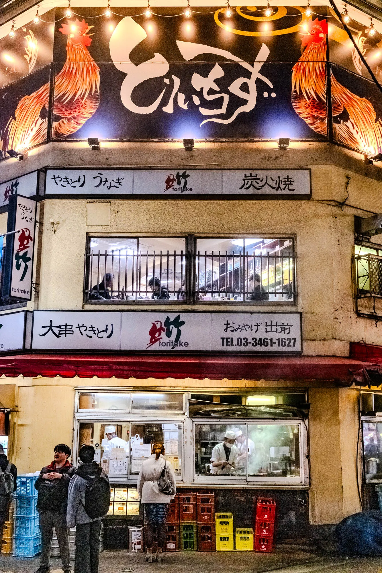 The image shows a Japanese restaurant. The front of the building is lit up with string lights. Above the doorway, there is a sign with a rooster on it. The sign says "e" and has Japanese characters beneath it.  Beneath the sign, there is a row of signs with the name of the restaurant and the different types of food they serve, including yakitori (grilled chicken skewers), eel, and "char-grilled". The windows are covered with metal bars. There is a large window in the middle of the building that shows the inside of the restaurant. There is a man wearing a white hat in the window preparing food. There are three customers outside the restaurant waiting. 

There is a sign at the bottom of the building that says "Obuge Delivery" and has a phone number.