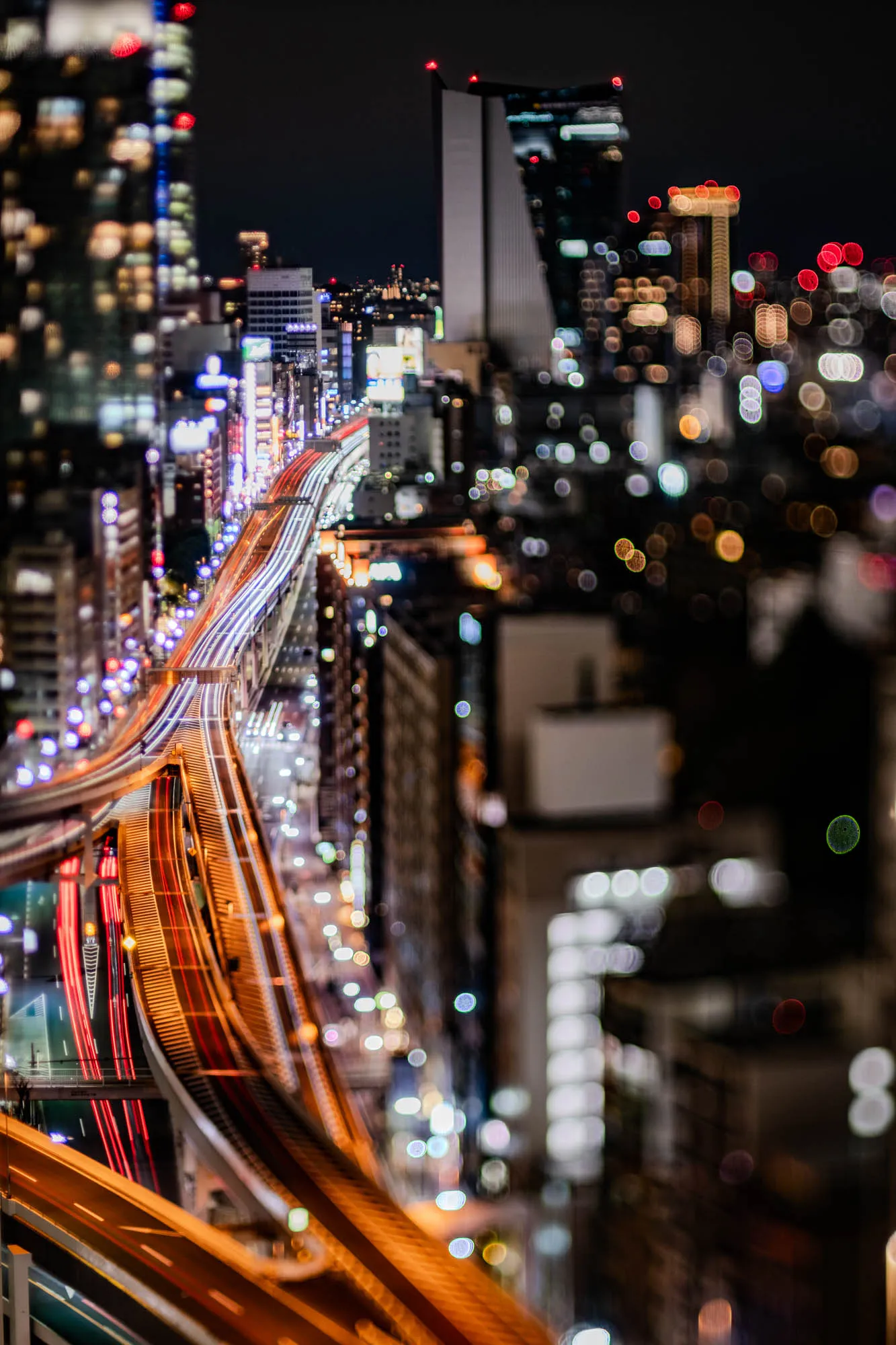 The image is a night time view of a city, seen from a high vantage point. The primary focus is on the highway, which is several lanes wide and appears to be elevated, with many cars driving on it. The car headlights appear as long streaks of white light, and the taillights are long streaks of red light. The highway is surrounded by buildings that are blurred in the background, and it appears that this highway system is quite complex, with several ramps and overpasses. The city lights blur into a sea of different colours, creating a hazy, sparkling effect in the background.  The city appears to be bustling with activity. The image is taken at night, with the only source of light being the city lights and the car headlights. The overall atmosphere is one of energy and movement.