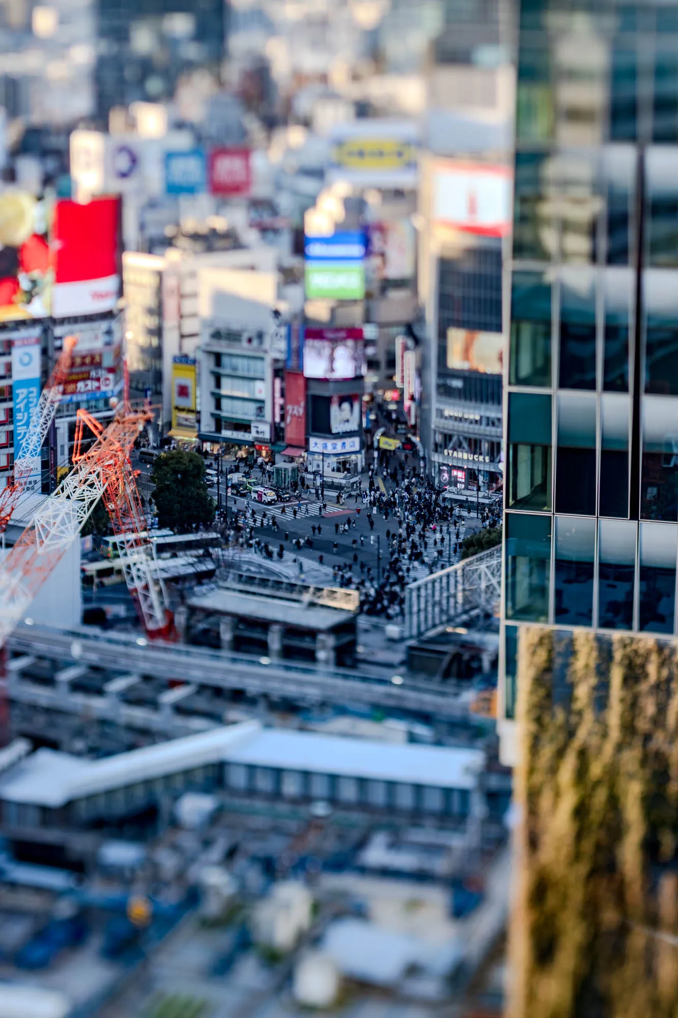 This is an aerial view of a city.  The image is out of focus, but it appears to be a cityscape with many buildings, a large crane, and a busy street with pedestrians.  You can see a Starbucks sign on one of the buildings. To the right of the image, there is a building that has a leafy green covering on the outside.  The city appears to be a bustling, densely populated area.