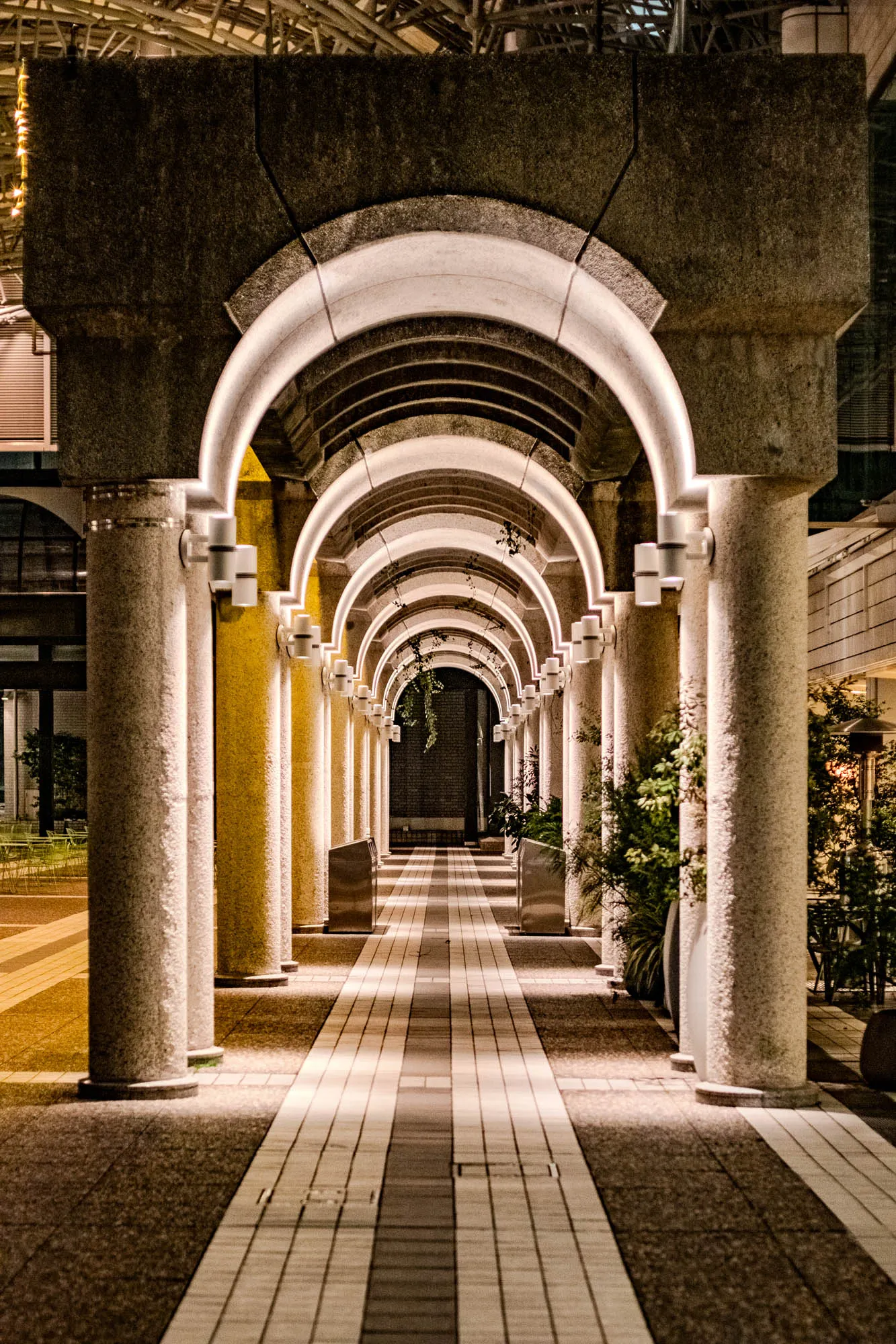 The image depicts a long, narrow walkway lined with evenly spaced stone pillars. Each pillar has a rounded archway on top, creating a series of interconnected arches that extend the length of the walkway.  The walkway is made of light-colored stone tiles, creating a  striking contrast with the darker stone of the pillars. The arches are illuminated by soft, white lights emanating from small fixtures mounted on the pillars.  There are some green plants growing along the sides of the walkway, adding a touch of natural greenery to the otherwise urban setting. The perspective of the image is such that the arches appear to recede into the distance, creating a sense of depth and mystery. The overall mood is one of tranquility and order, with the symmetry of the arches and the even spacing of the pillars creating a sense of harmony.  This image is a classic example of architectural design. 
