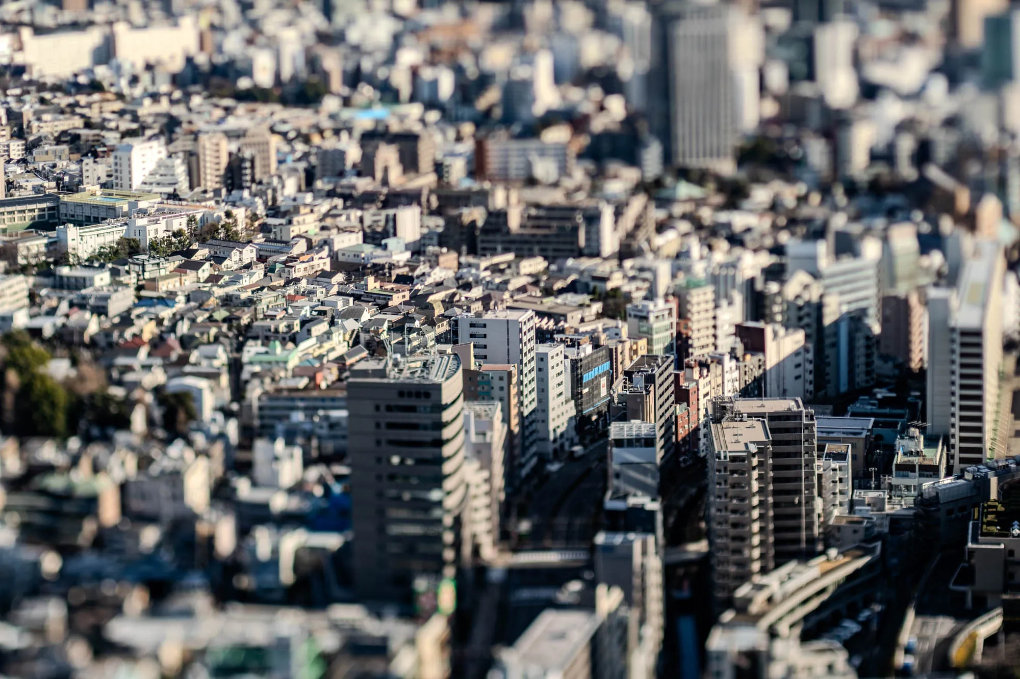 The image shows a cityscape from a high vantage point, with the buildings appearing small and blurry, creating a miniature effect. The majority of the scene is made up of closely clustered buildings of various heights and shapes, most appearing in shades of gray and brown. The buildings in the foreground are slightly clearer than those in the background. The effect suggests a dense, urban environment. The image likely captures a city like Tokyo, known for its many buildings and crowded spaces.