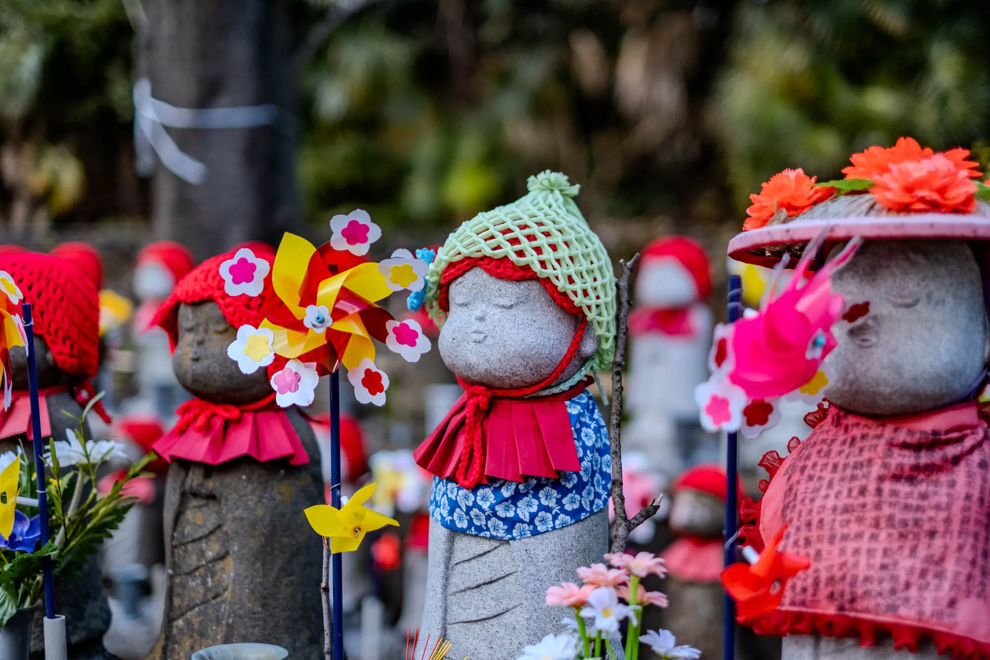 This image shows a group of small stone statues. The statues are all in the shape of children and are dressed in colorful clothes and hats. The statues are surrounded by flowers and other decorations. In the foreground, a statue wears a light green crocheted hat and a red and blue outfit. They also have a pink, yellow, and red pinwheel in front of them. Behind the statues, a tree trunk can be seen out of focus.