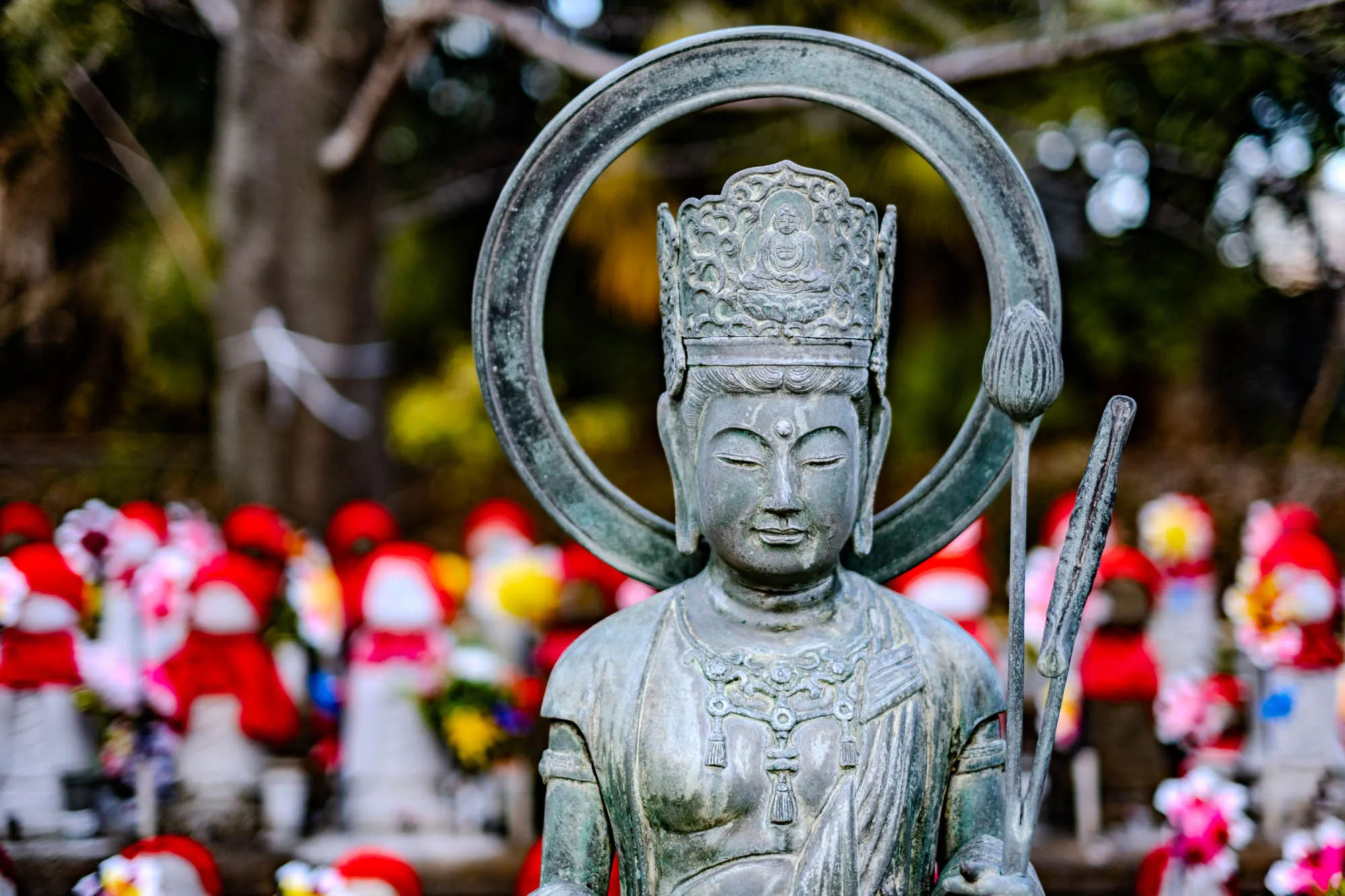 The image shows a close-up of a weathered bronze statue of a person. They have a calm expression on her face with her eyes closed. They are wearing a large crown with intricate details and a beaded necklace that hangs down her chest. They are holding a large staff in their left hand, and a smaller staff in their right hand. They are surrounded by an out-of-focus halo and is standing in front of other blurry statues, many of which are covered in red cloth and flowers. The background is blurred, suggesting an outdoor location with trees and greenery. 
