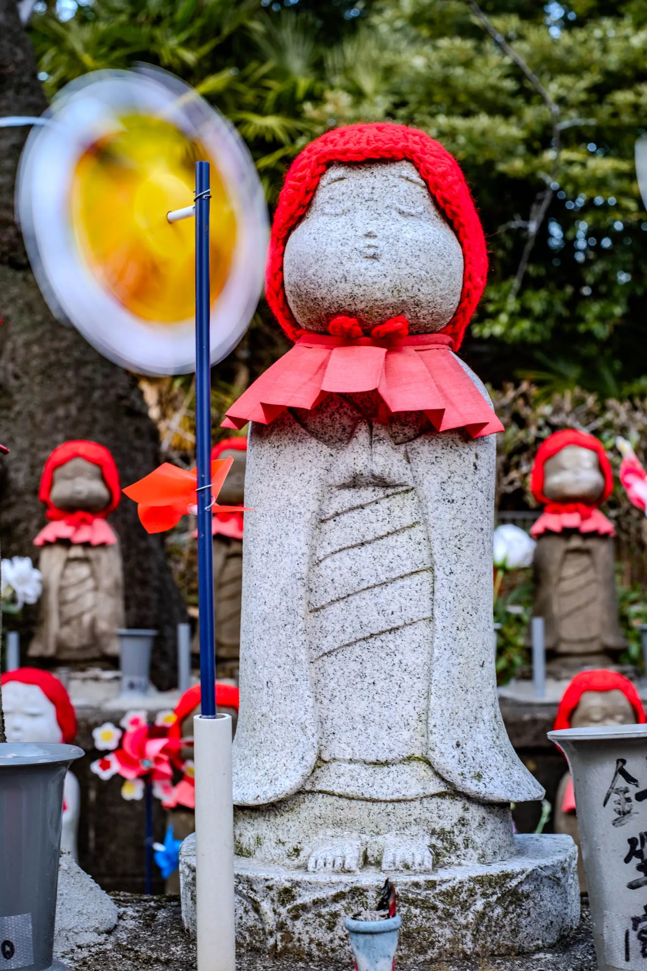 The image shows a stone statue of a person with a red knitted hat and a red cloth around their neck. The statue is standing on a stone base and has a white, gray and slightly green speckled finish. The statue has their hands clasped in front of them. The statue is standing in front of a blur of a yellow and white pinwheel that is spinning. To the right of the statue, there are two other stone statues with red hats, one with a red cloth on its neck and the other with a red cloth wrapped around its torso. There are other statues that are partially visible.  Behind the statues, there are some trees with green leaves. To the left of the statue, there is a partially visible grey planter. There is a white and blue stick attached to the grey planter. The ground appears to be grey, rocky, and uneven.  In the bottom right corner of the image, there is a grey container. The container is labeled with black writing that is illegible. There is a grey container in the background with writing in black. The grey container and the statue in front of it are in focus while the background and the pinwheel are blurred.