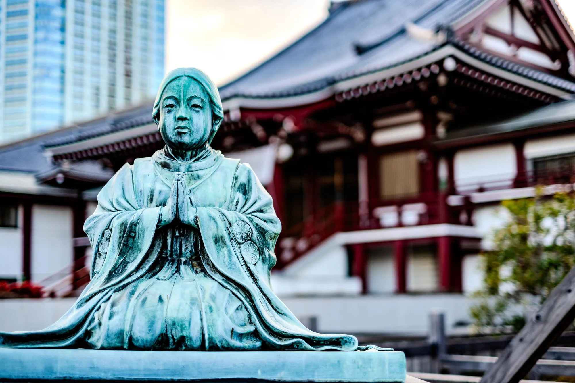 The image shows a bronze statue of a woman kneeling with her hands clasped in prayer. The statue is weathered and has a greenish-blue patina. She is wearing a long robe with a large collar. The statue is in front of a blurry background of a traditional Japanese temple with a red roof and a white building. There is also a tall, modern building in the background. The temple has a tiered,  multi-level roof with distinctive angles. There is some foliage visible on the right side of the image.  There is a dark wooden fence behind the statue.