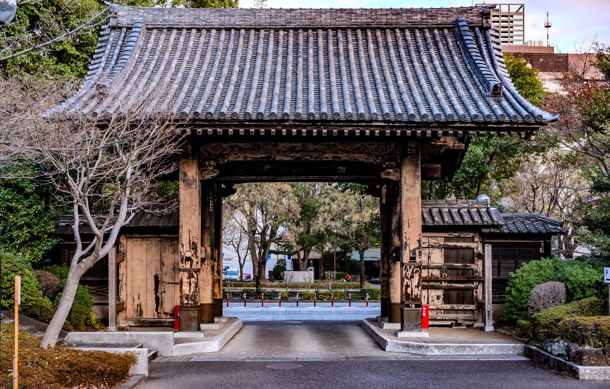 The image shows a traditional Japanese gate, a Torii, made of wood and gray tile, with a curved roof. The gate is standing open and there are two thick wooden posts on each side of the entrance. The gate stands in front of a paved road, which is lined with trees on both sides. The road leads to a street with a small statue and some pedestrians. Behind the gate, there are some green bushes and trees and a building with a gray tiled roof. You can also see a sign for parking on the right.  The wood of the gate looks weathered and aged. On the left side of the image, a bare tree with thin branches grows in the dirt. The gate is a beautiful example of Japanese architecture.