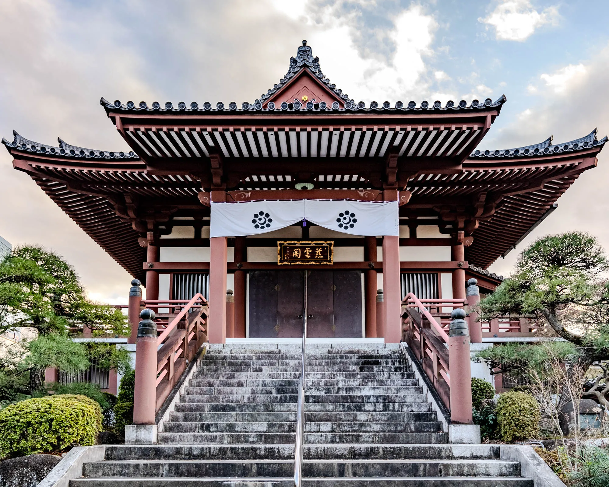 The image shows a traditional Japanese temple with a large, intricately-designed roof. The roof is made of red wood with a black tiled overhang. It is supported by a series of wooden beams, which create a horizontal, striped pattern on the underside of the roof. The temple has a large, white banner hanging from the roof above the entrance. The banner has black circles in each corner and a design in the middle with black circles around it.  Below the banner is a wooden sign with gold writing on it.  The sign hangs above a set of double wooden doors.  The doors are dark brown with a vertical, wood-slat design. In front of the doors is a set of stairs made of stone. The steps are uneven and have a gray, mottled surface. On either side of the stairs are red wooden railings with black, orb-shaped posts. The rails have an elaborate design on them.  There are trees on the left and right of the temple and a small bush to the left of the stairs. The sky above the temple is a pale blue with a few white clouds.