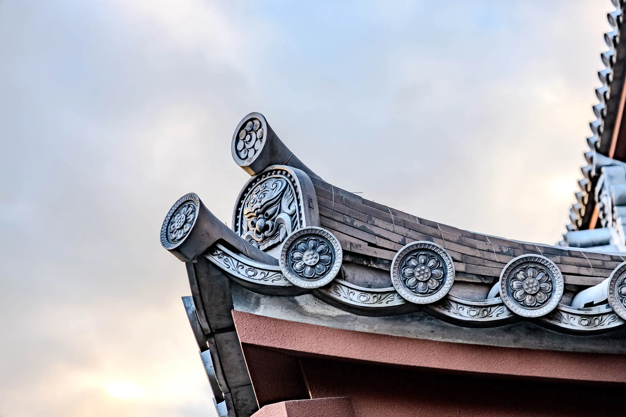 The image shows a close-up of a traditional Japanese roof. The roof is made of dark gray tiles and has a curved shape. The roofline is adorned with intricate, decorative elements, including a series of circular medallions, each featuring a stylized floral design.  The medallions are positioned along the roofline, with the top one being the most ornate. The medallions are made of a silver material that contrasts sharply with the dark gray tiles.  The roof is also decorated with a series of small, triangular shapes that run along the edge. The background is a bright sky with a few clouds.  There is a large, decorative element that is shaped like a horn or a beak, and has a face carved into it, near the top of the image. The decorative element is positioned at the peak of the roof. It is made of a similar silver material and is decorated with intricate carvings. The image is taken from a low angle, giving the viewer a good view of the roof's ornate details. The image evokes a sense of beauty, craftsmanship, and tradition. 
